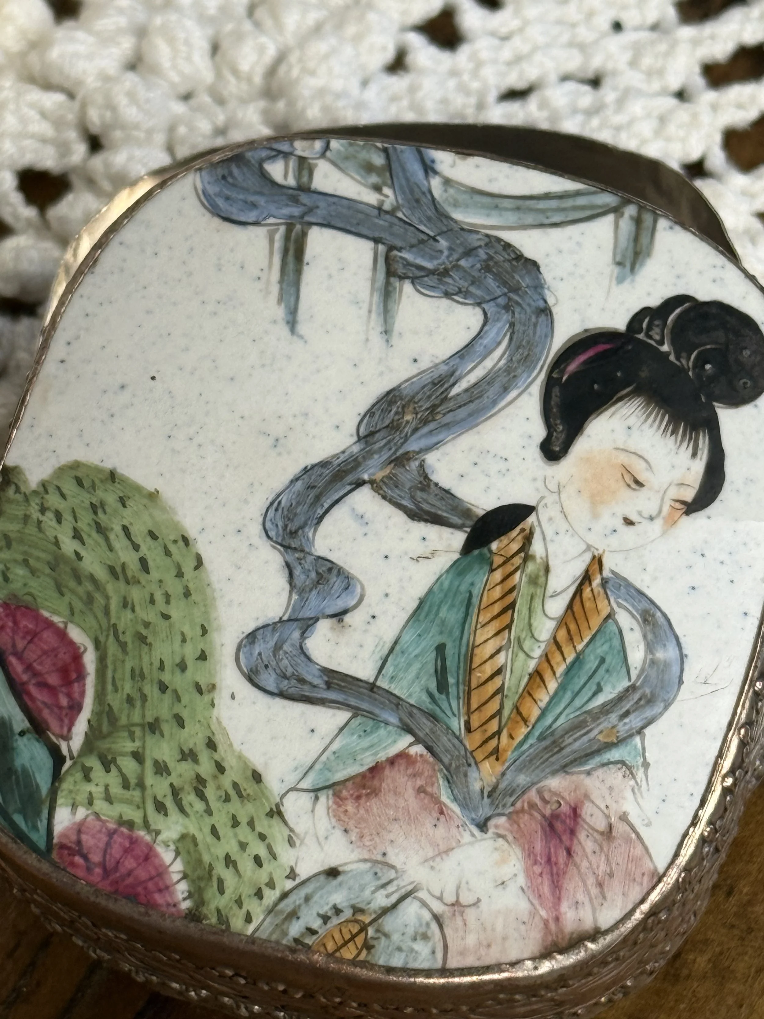 Angled close-up of hand-painted porcelain lid showing traditional Asian figure, bamboo landscape, pastel robe details, and painterly brushwork on vintage ceramic surface