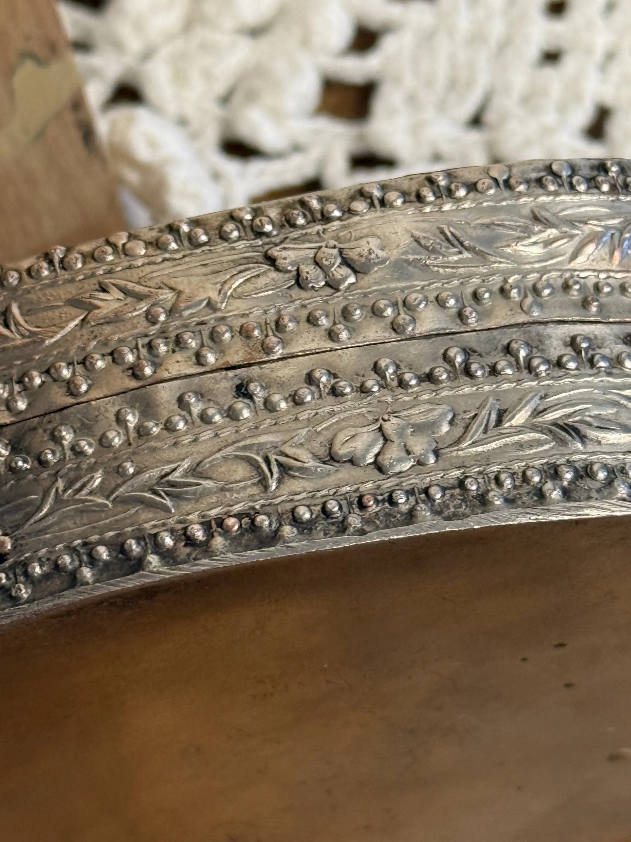 Macro detail of repoussé silver-toned metalwork with floral and vine motifs, raised bead borders, and aged vintage patina on decorative trinket box base