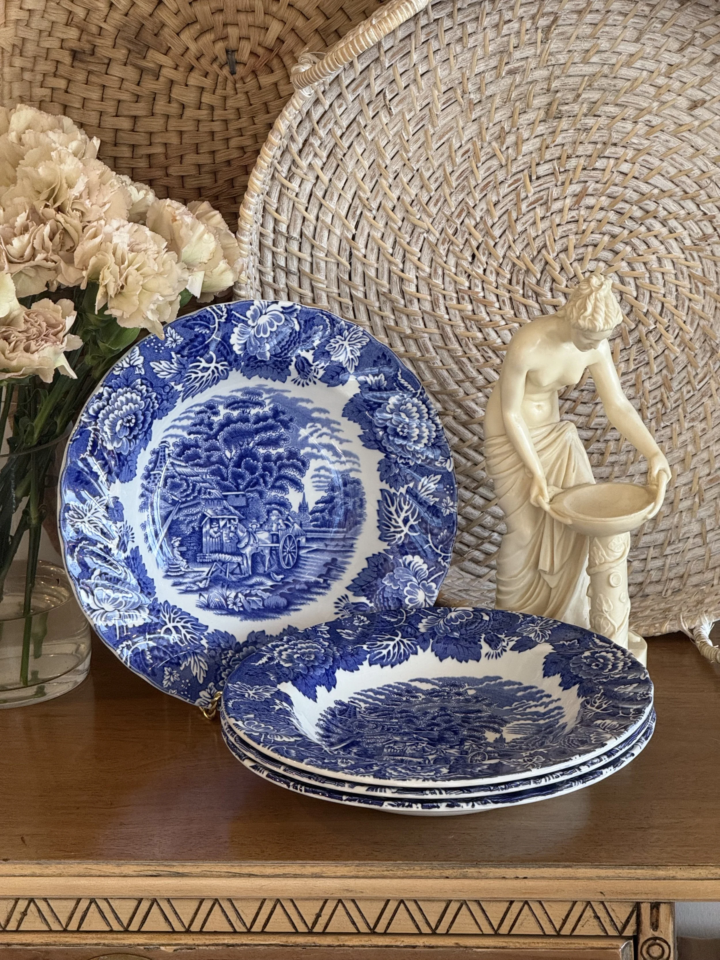 a set of stacked Vintage blue transferware plates on a wood table surrounded by a white wicker tray, soft blooming flowers and an alabaster statue figurine.