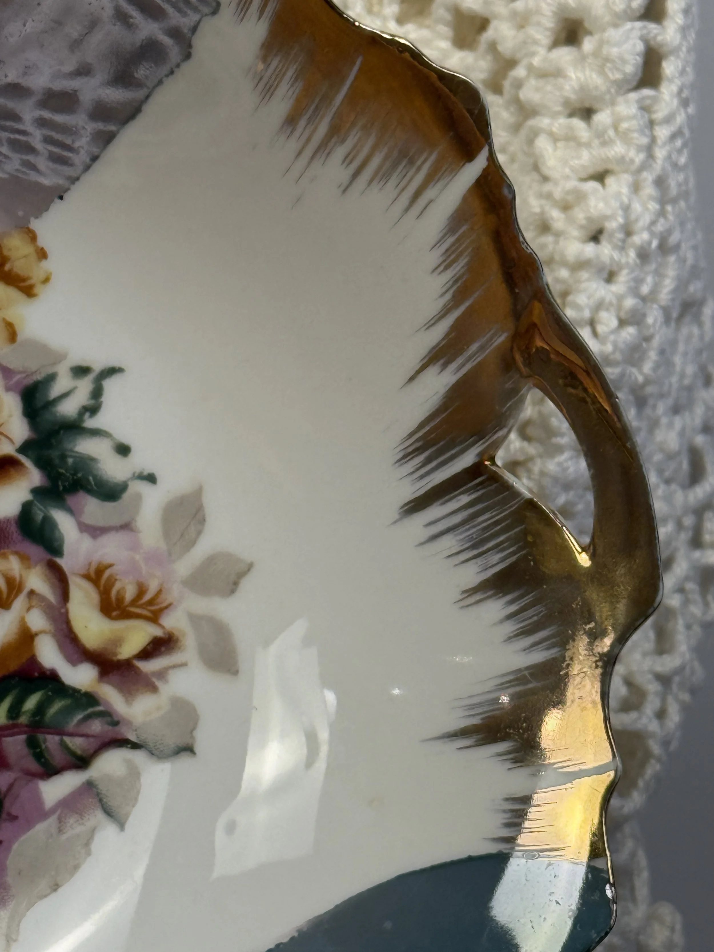 Close-up detail of scalloped porcelain plate rim showing expressive grey-blue brushstroke glaze, warm amber-gold edge accents, and fine gold trim on vintage floral plate