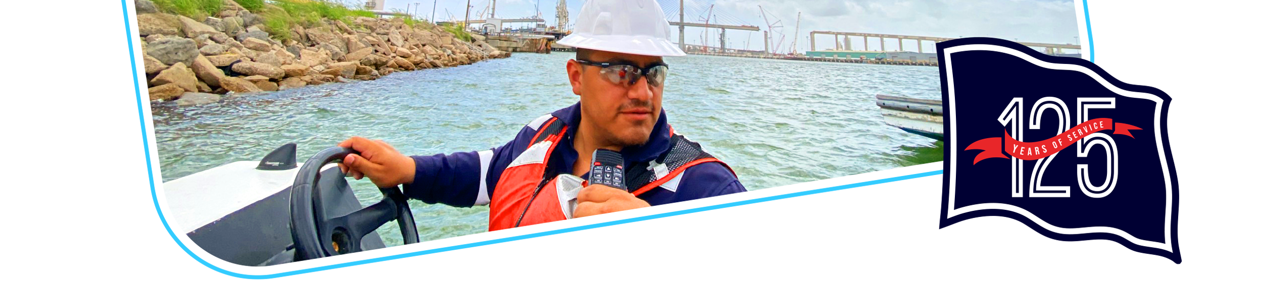 A man in safety gear and sunglasses holding a radio on a boat in a harbor with rocks and construction cranes in the background. There is a graphic of a flag with the number 125 and a banner that says 125 years of service.