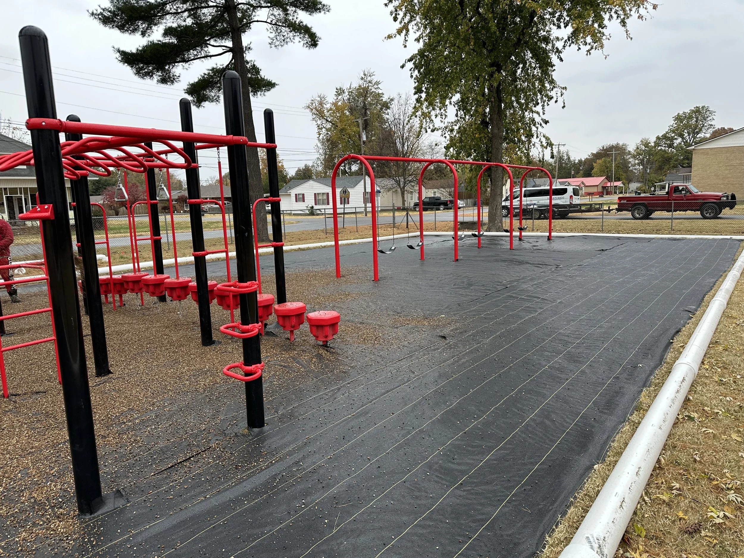 Empty playground with black and red climbing structures and swings, partially covered with black rubber safety surfacing, and a white pipe along the edge in a residential area with trees and parked cars in the background.