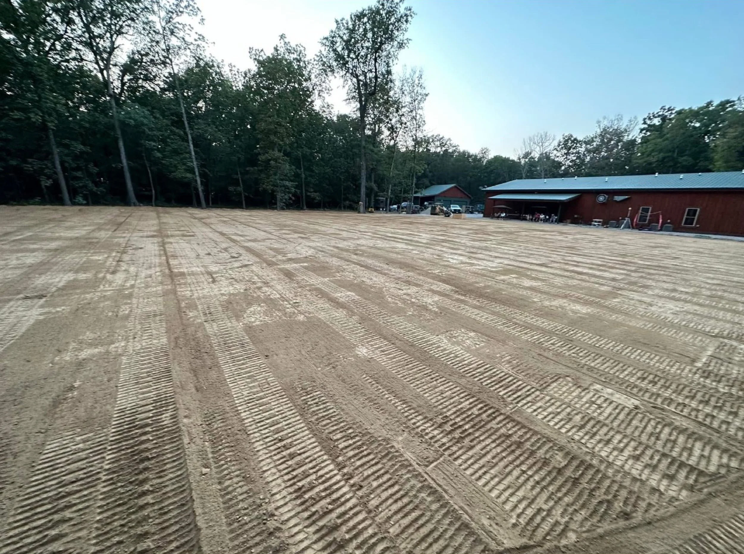 A large cleared dirt area with tire tracks, bordered by trees and a red barn-style building with machinery and people in the distance.