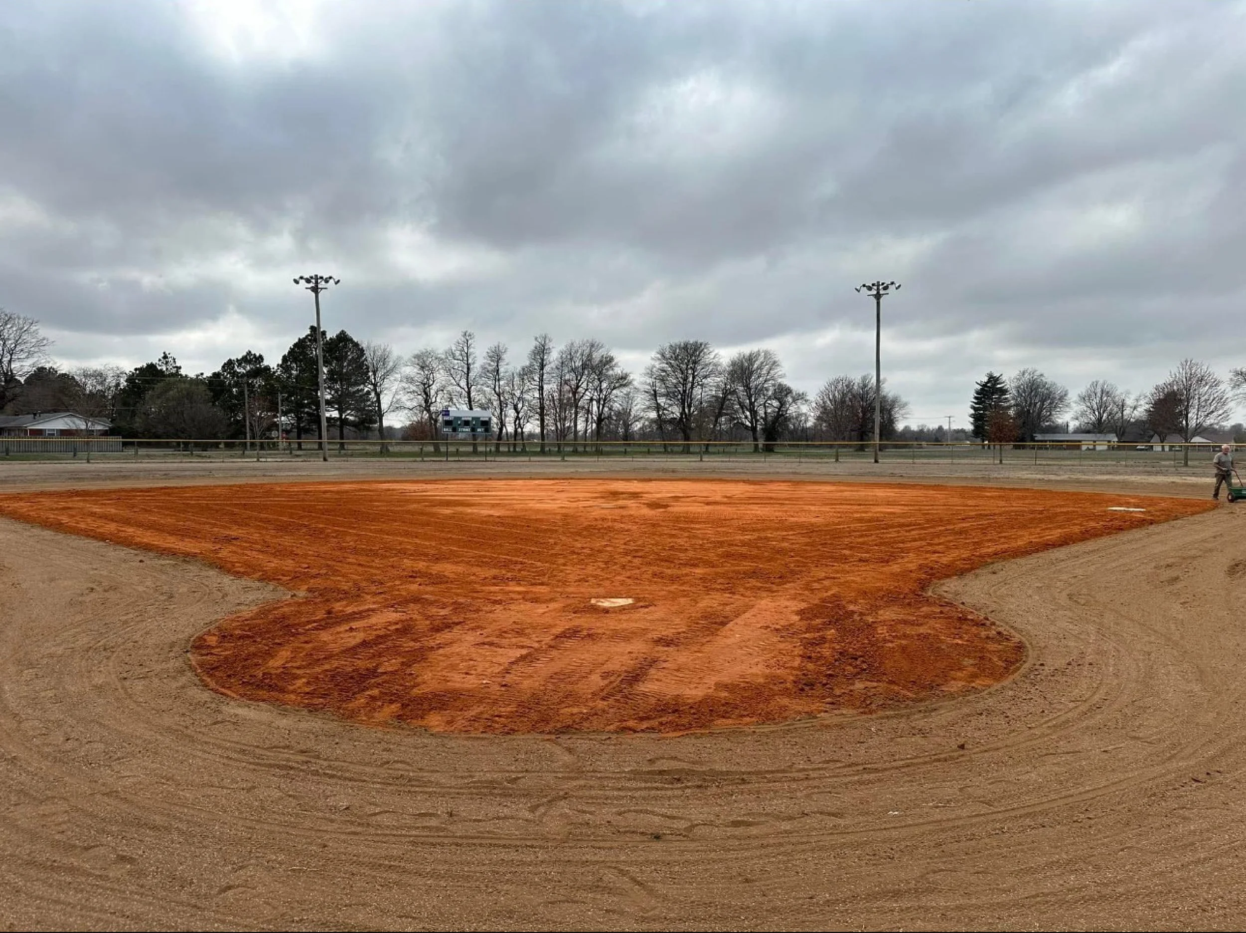 Empty baseball field with freshly groomed dirt and grass under cloudy skies, with trees and bleachers in the background.