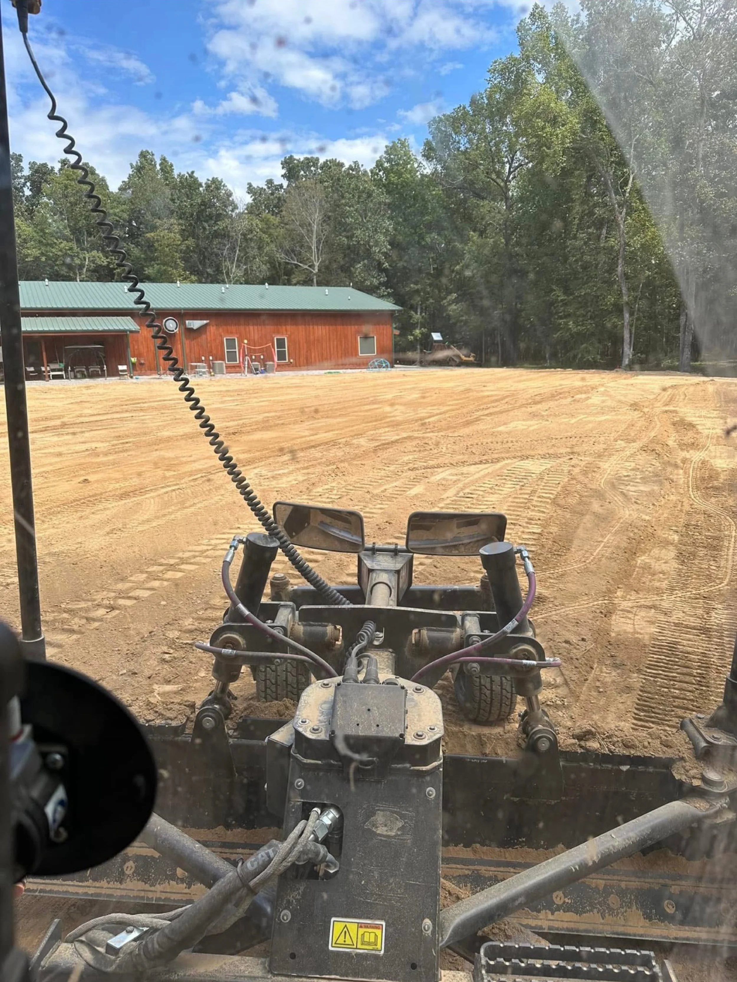 Inside view of a bulldozer working on a dirt field with a wooden building and trees in the background.