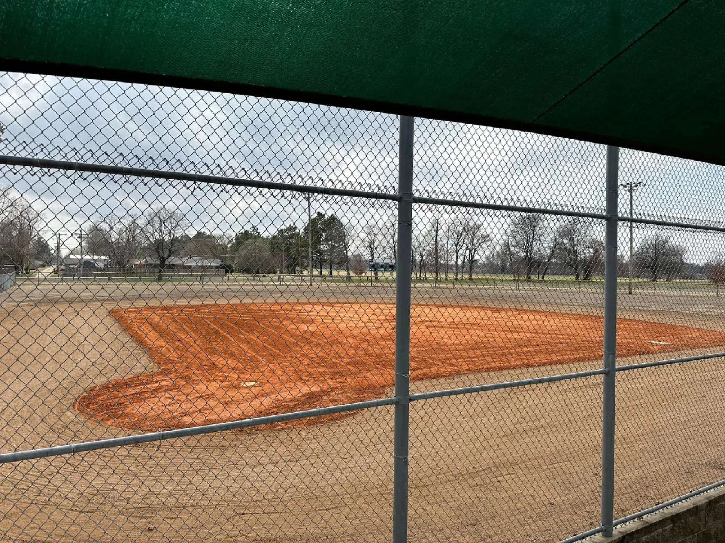 A baseball field under construction with a dirt infield and a simple chain-link fence surrounding it, viewed from behind a green shade cover.