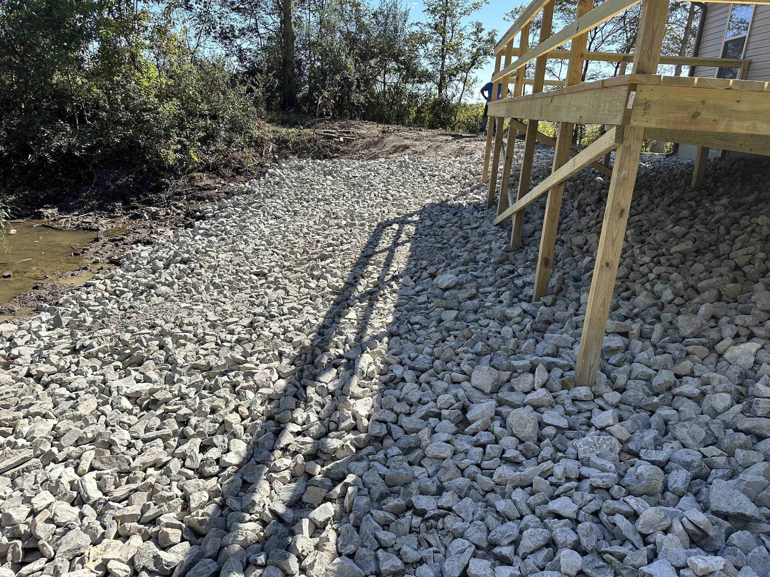 A construction site showing a wooden staircase structure being installed on a gravel slope next to a house, with trees and bushes in the background.