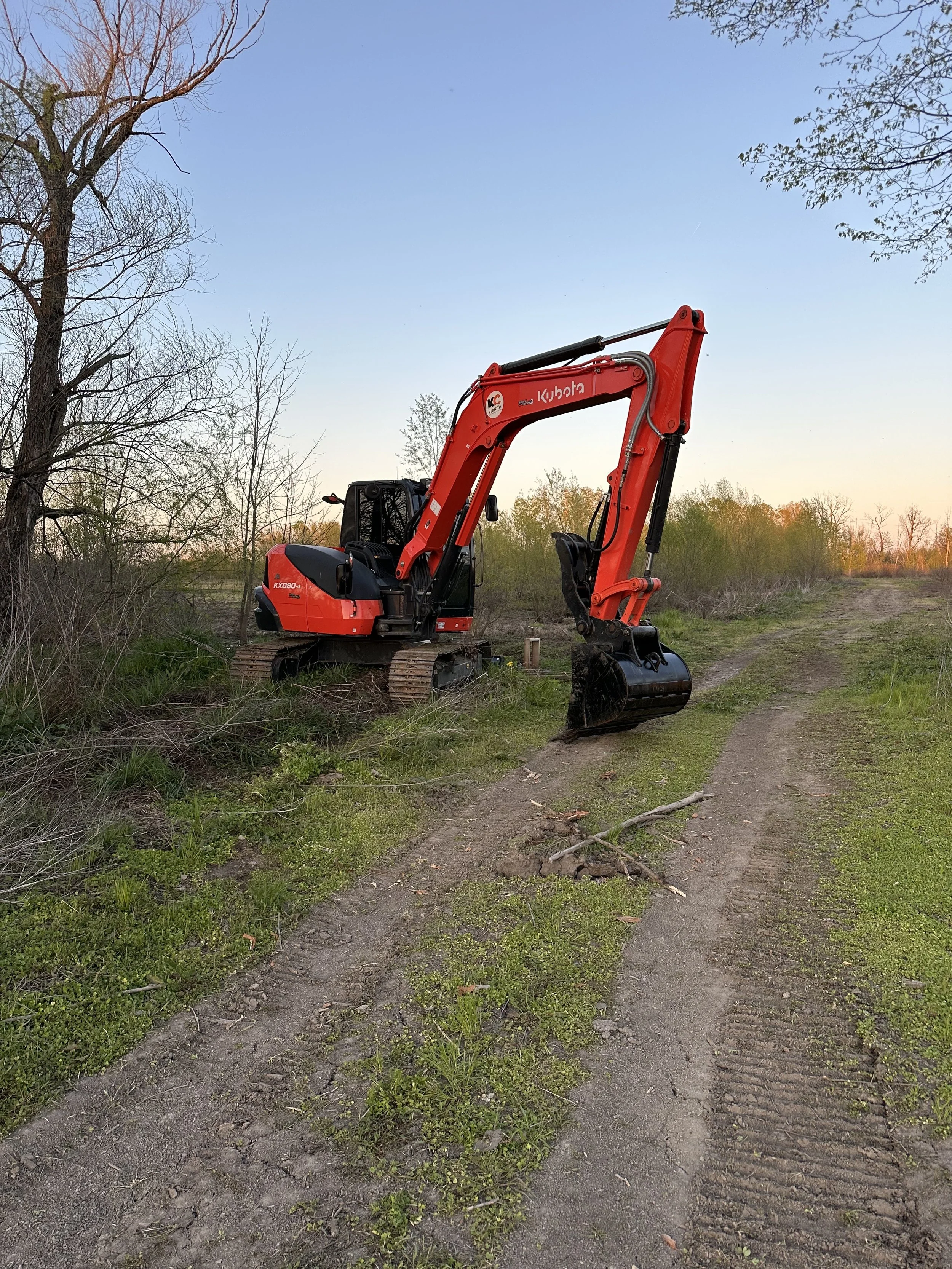 A small red Kubota excavator situated on a dirt trail in a natural outdoor setting with trees and greenery.