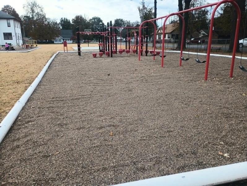 Empty outdoor playground with red swings and climbing structures, surrounded by a gravel surface and a white border, with houses and trees in the background.