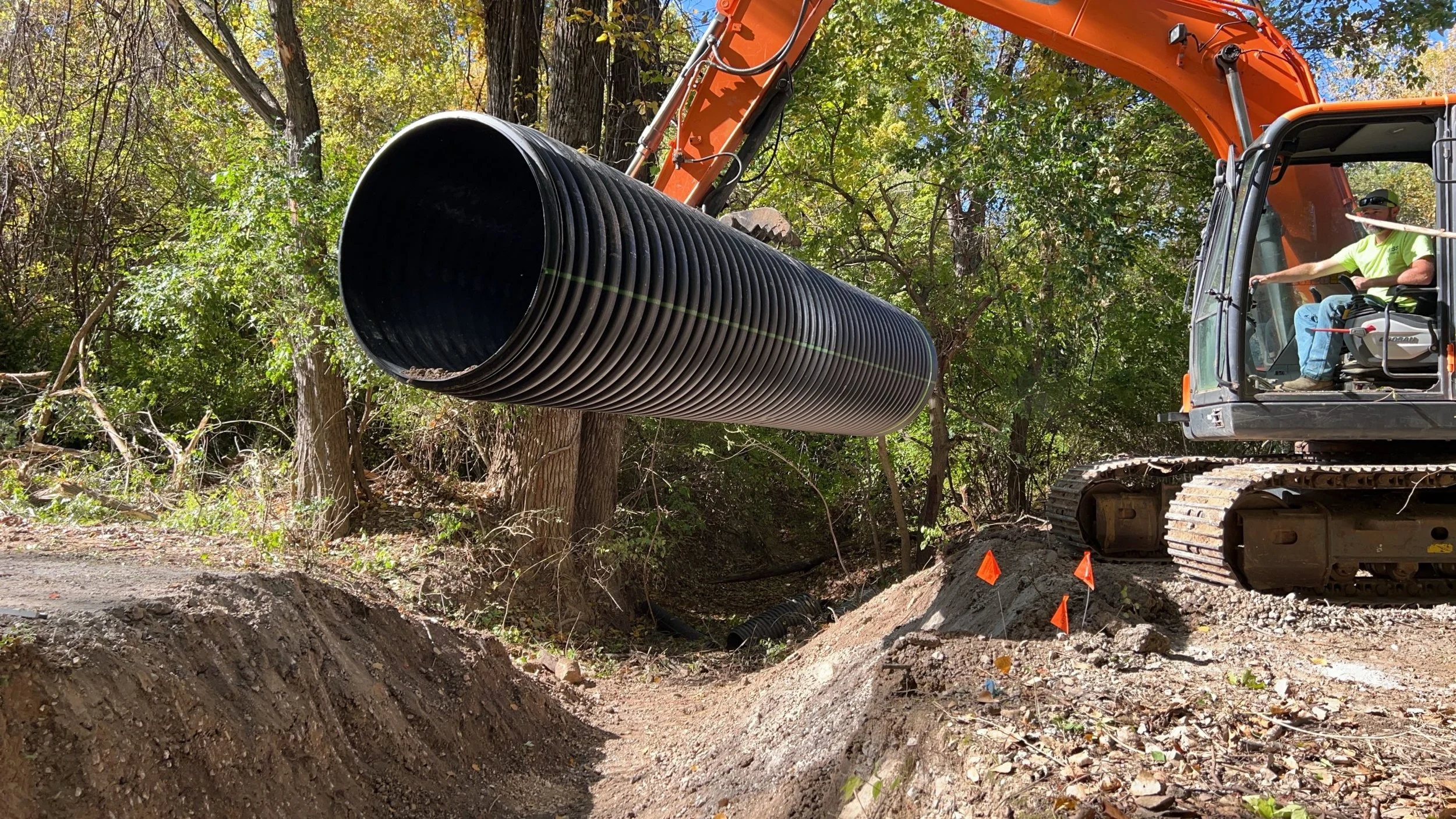 An excavator is installing a large black corrugated underground pipe among trees, with a worker operating the machine in a wooded outdoor area.