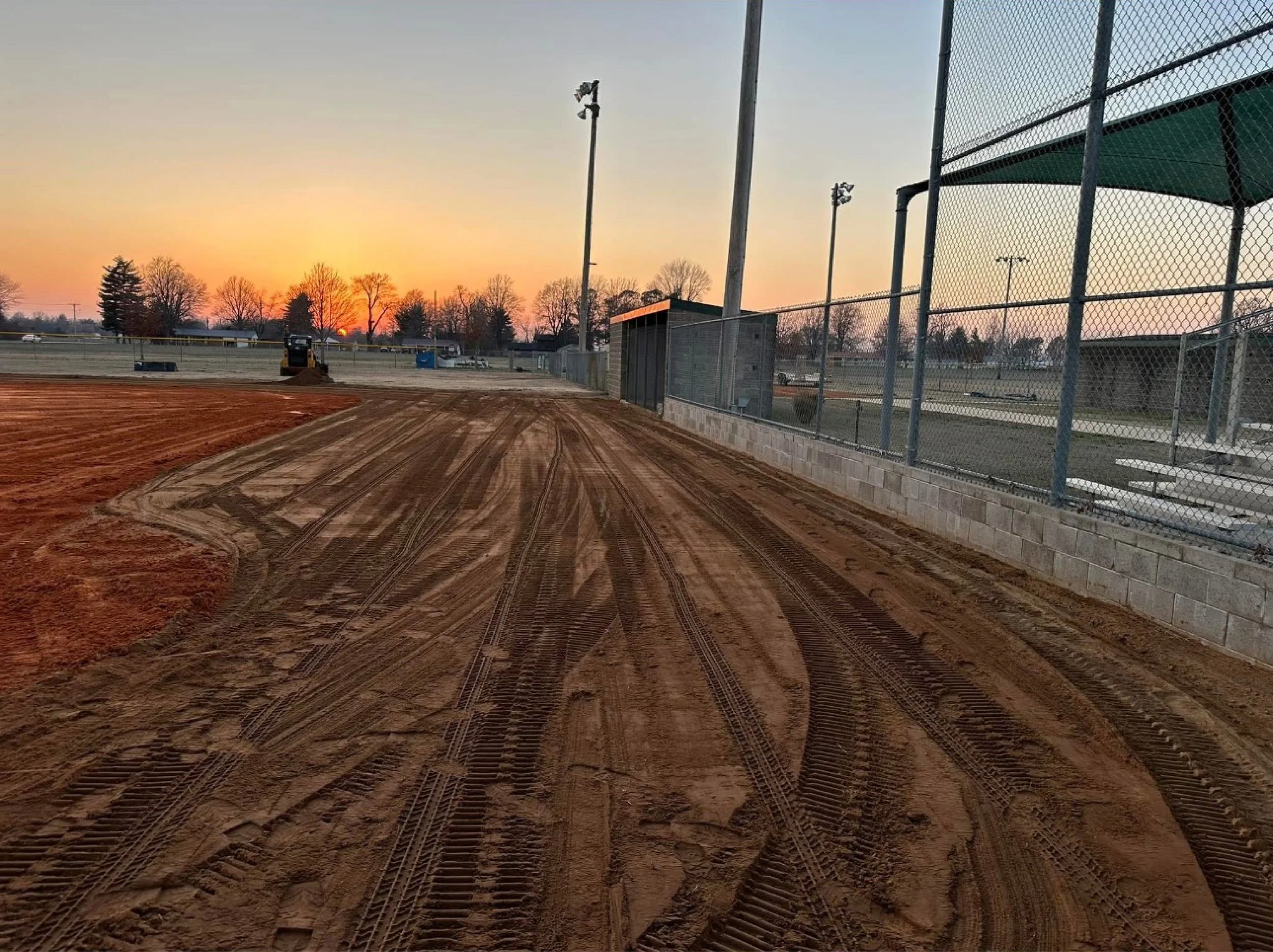 View of a baseball field under construction at sunset, with exposed dirt, tire marks, and a chain-link fence to the right.