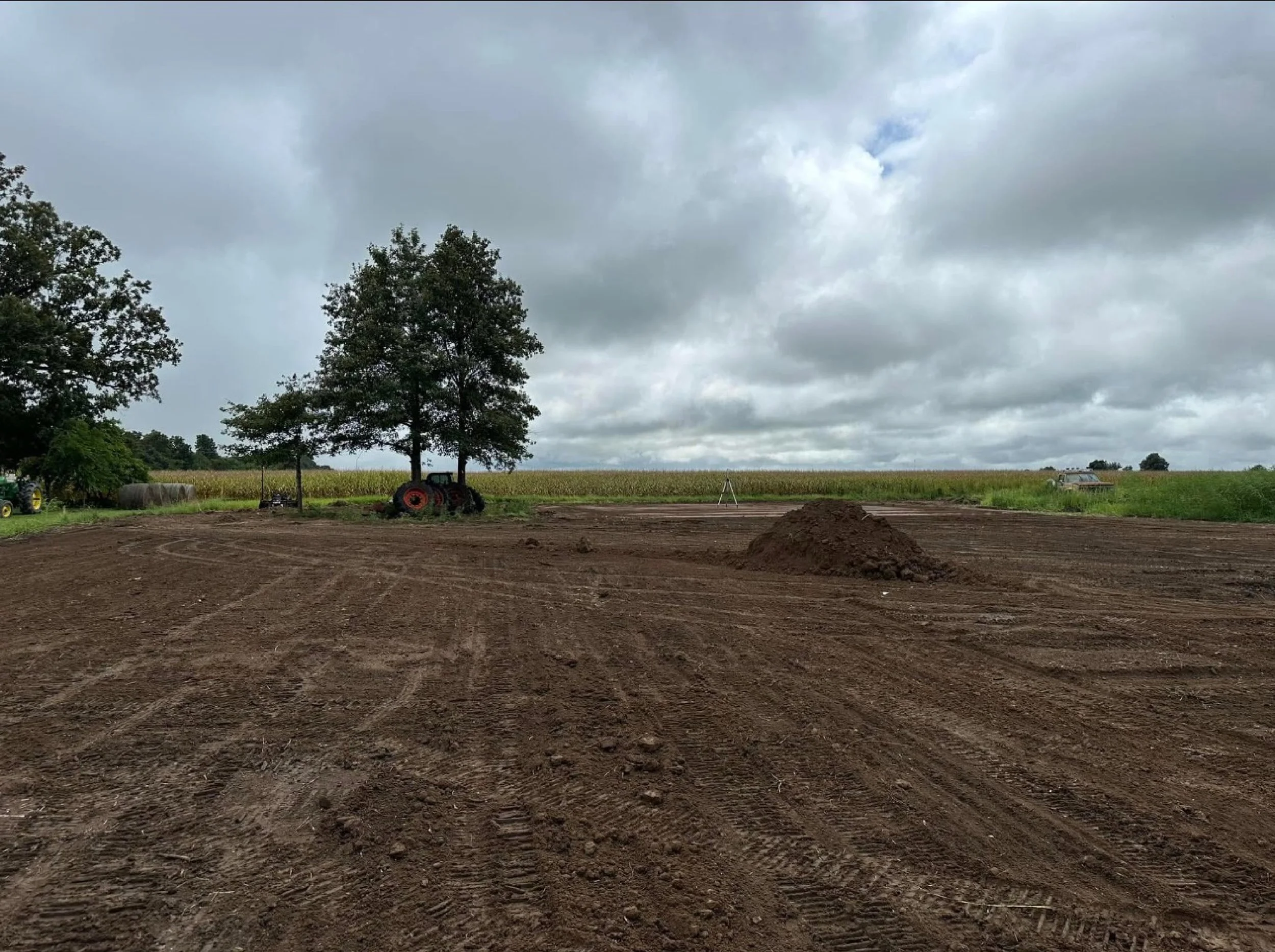 A farm field under a cloudy sky, with a large pile of dirt in the foreground and machinery, trees, and a crop field in the background.