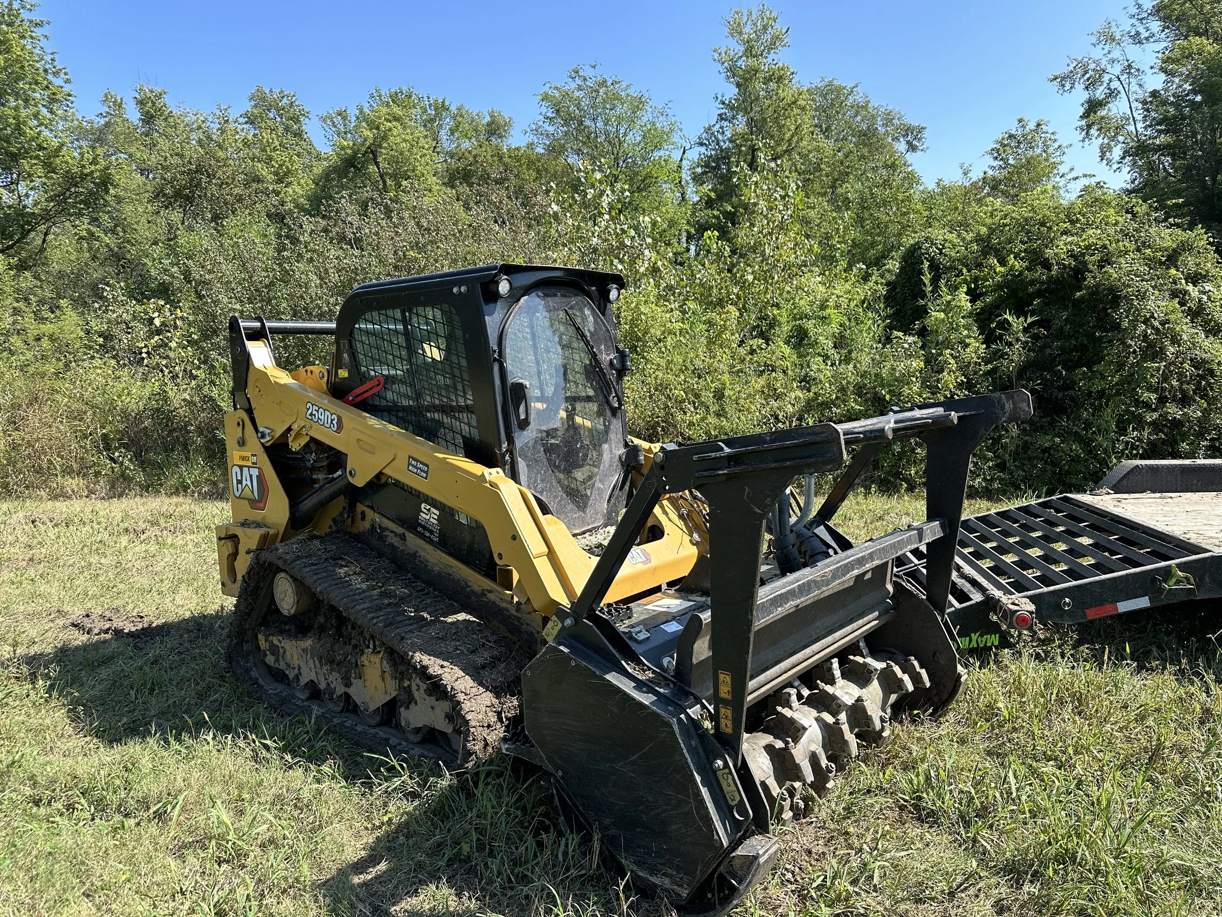 A Caterpillar compact track loader with a yellow and black color scheme parked on grassy terrain next to a trailer, with dense green trees and a clear blue sky in the background.