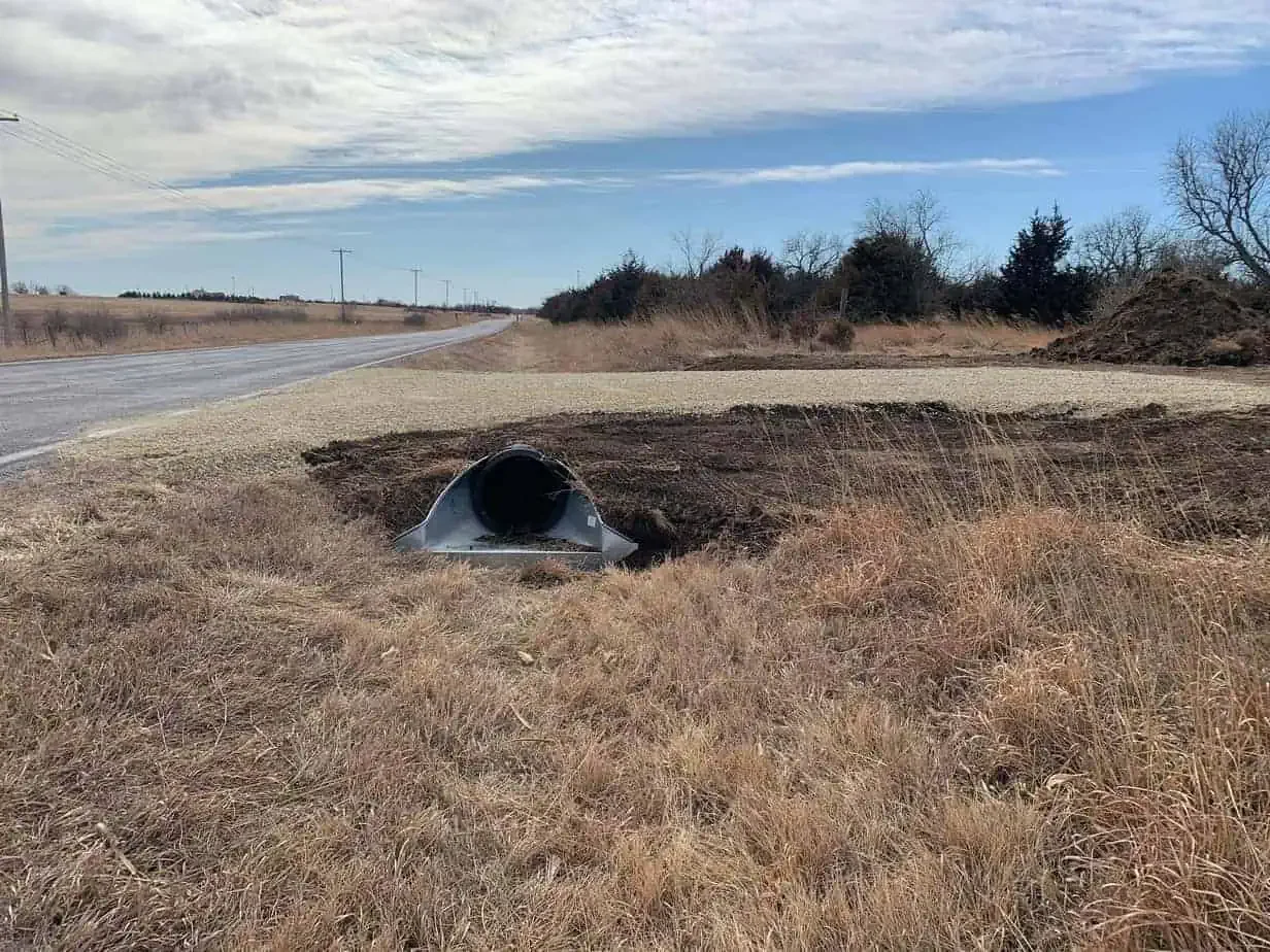 A metal culvert pipe on the ground beside a rural road, with dirt and dry grass around it, and utility poles in the distance under a partly cloudy sky.