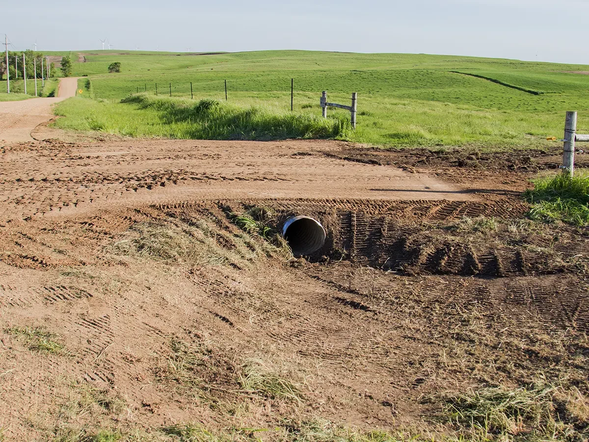 A rural dirt road with tire tracks and a large drainage pipe crossing beneath it, surrounded by green fields and rolling hills in the background.