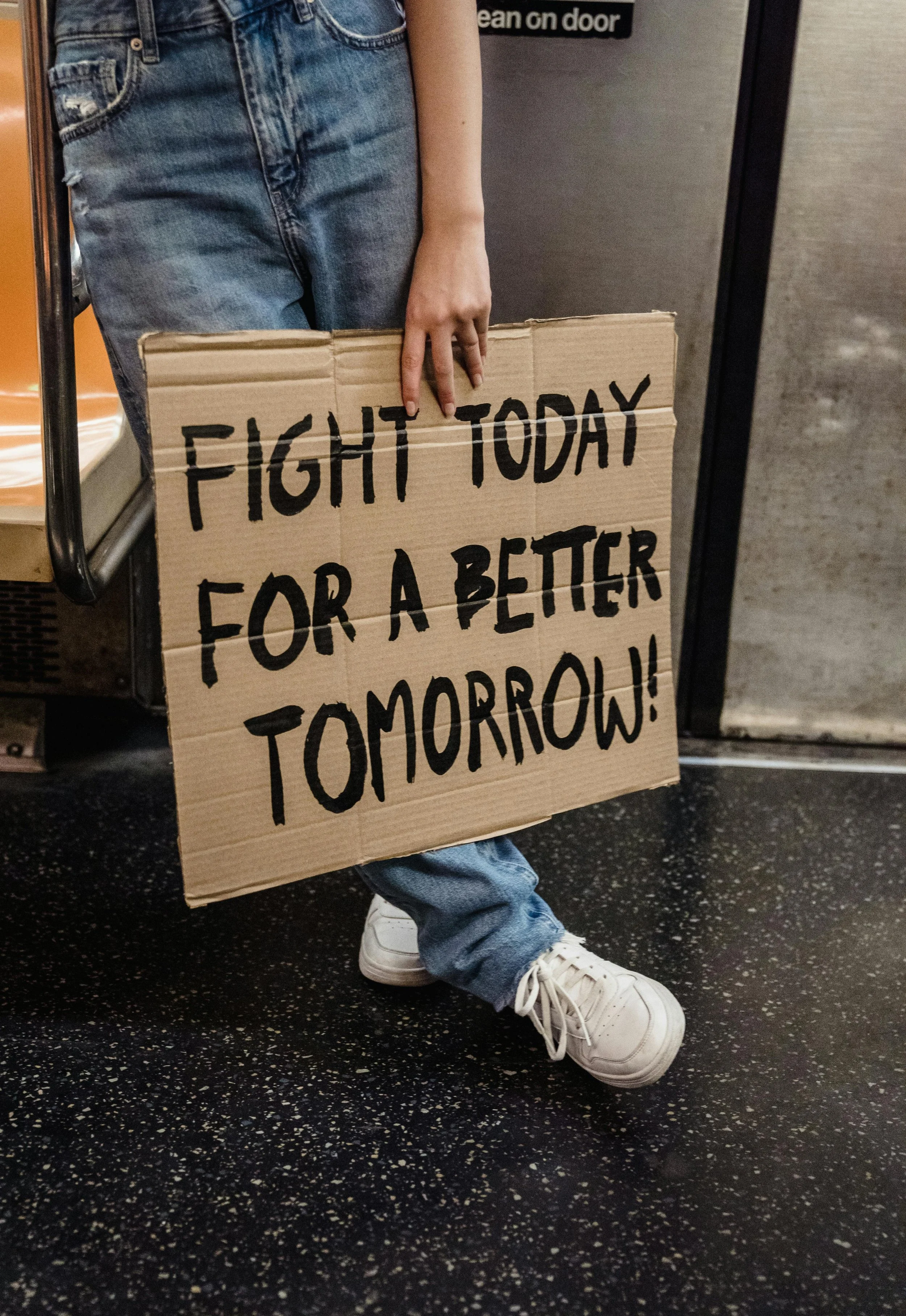 Person wearing jeans and white sneakers holding a cardboard sign that says 'FIGHT TODAY FOR A BETTER TOMORROW!'