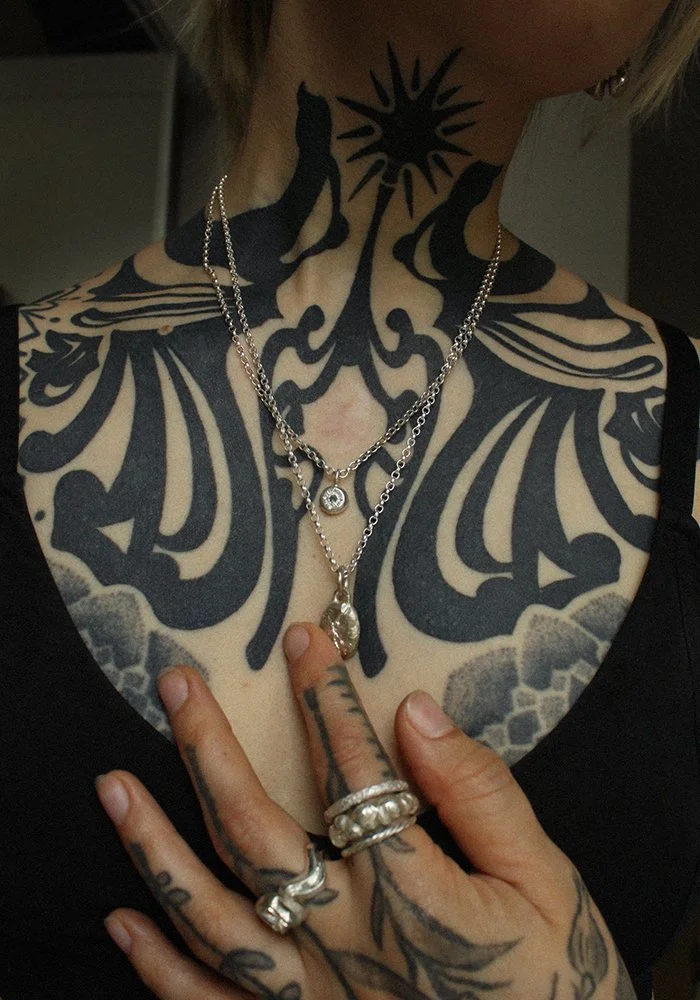 A person with extensive black tattoos on their neck, chest, and hand, wearing multiple silver necklaces and rings, and touching their tattooed chest with their tattooed hand.