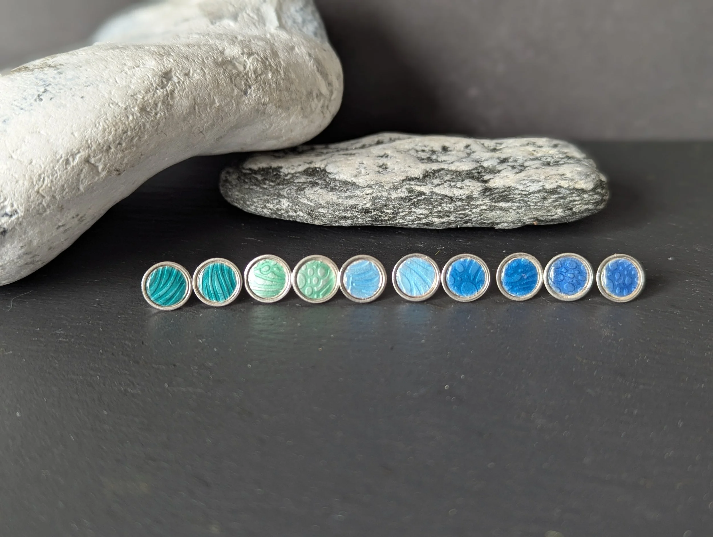 Set of ten small round earrings with blue and green abstract patterns, placed in a line on a black surface against two gray and white rocks.