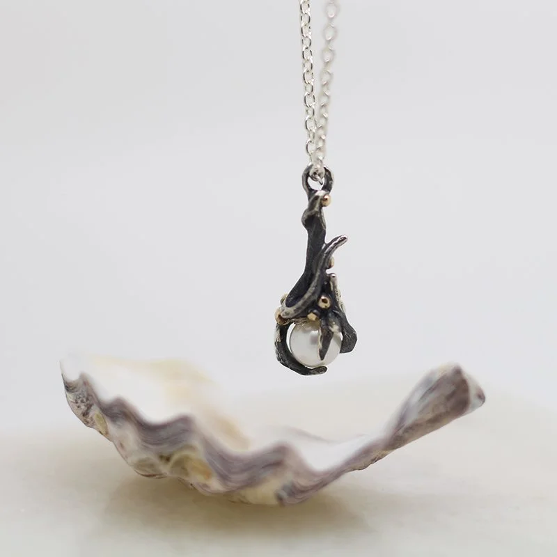 A necklace with a pendant shaped like a cat hanging from a branch, suspended above a shell on a white background.