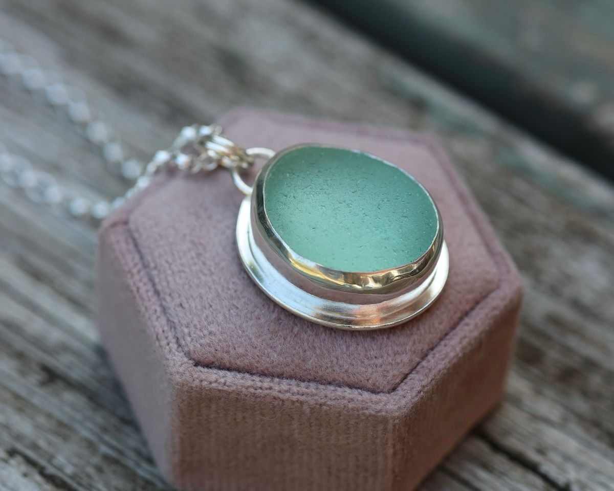 A silver oval ring with a large green gemstone, resting on a pink foam jewelry display pad, with a silver chain partially visible in the background, on a wooden surface.