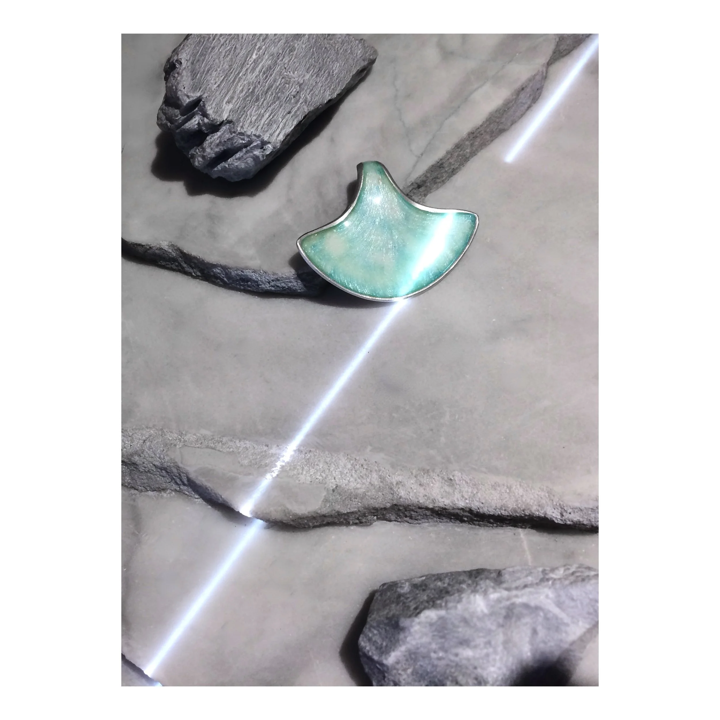 A seashell-shaped silver and turquoise jewelry piece resting on a gray stone surface surrounded by gray rocks.