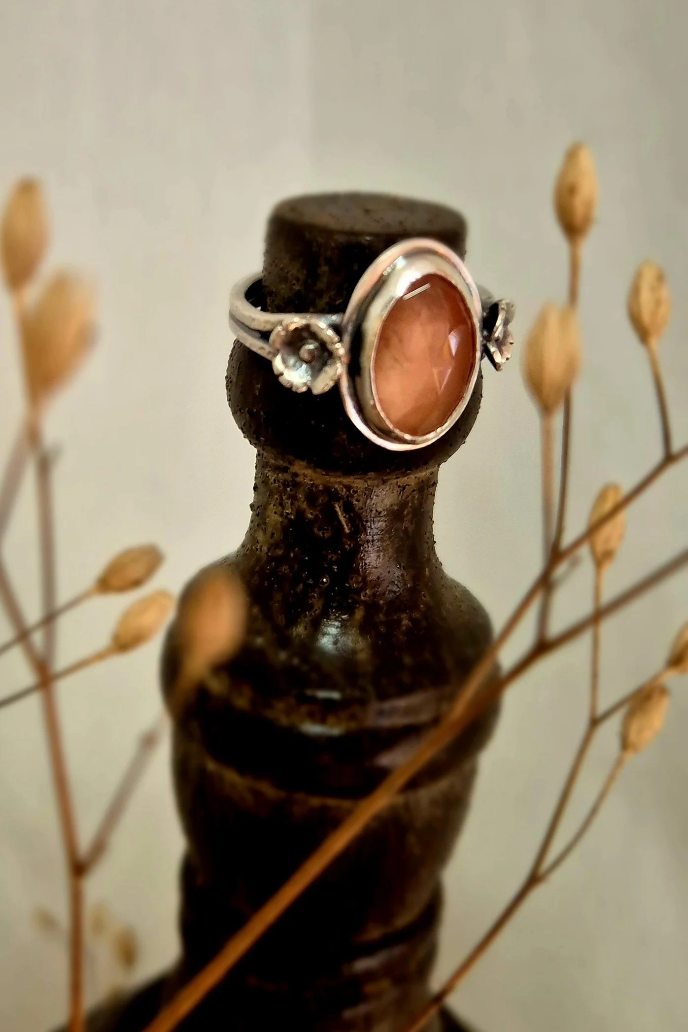 Ring with a large oval pink stone on a black lace jewelry holder, surrounded by dried plant stems.