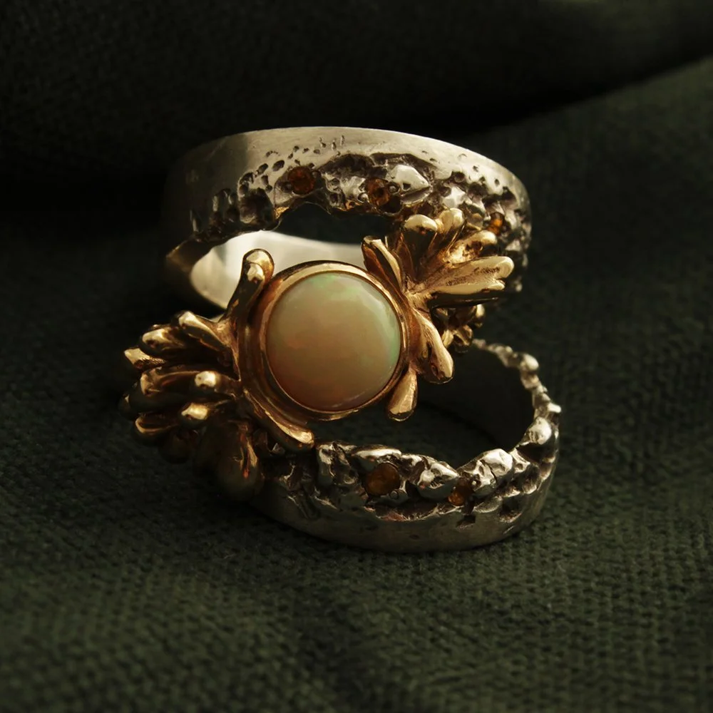 A pair of silver and rose gold rings with a central opal stone and leaf and branch designs, placed on dark fabric.
