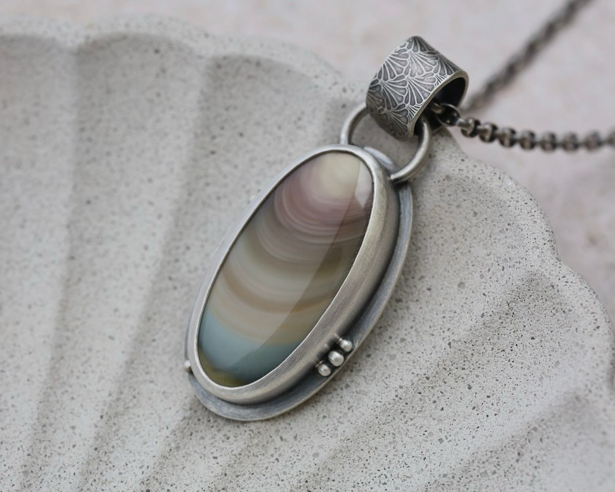 Close-up of a silver pendant necklace with an oval-shaped multicolored stone and a decorative bail, resting on a light textured surface.