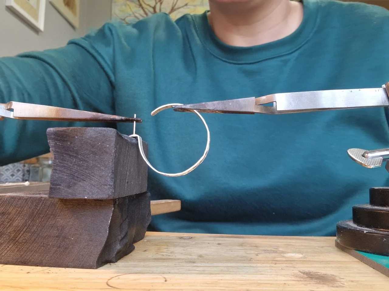 A person working on jewelry, using tools to shape a small metal ring with a black wooden block in the background.