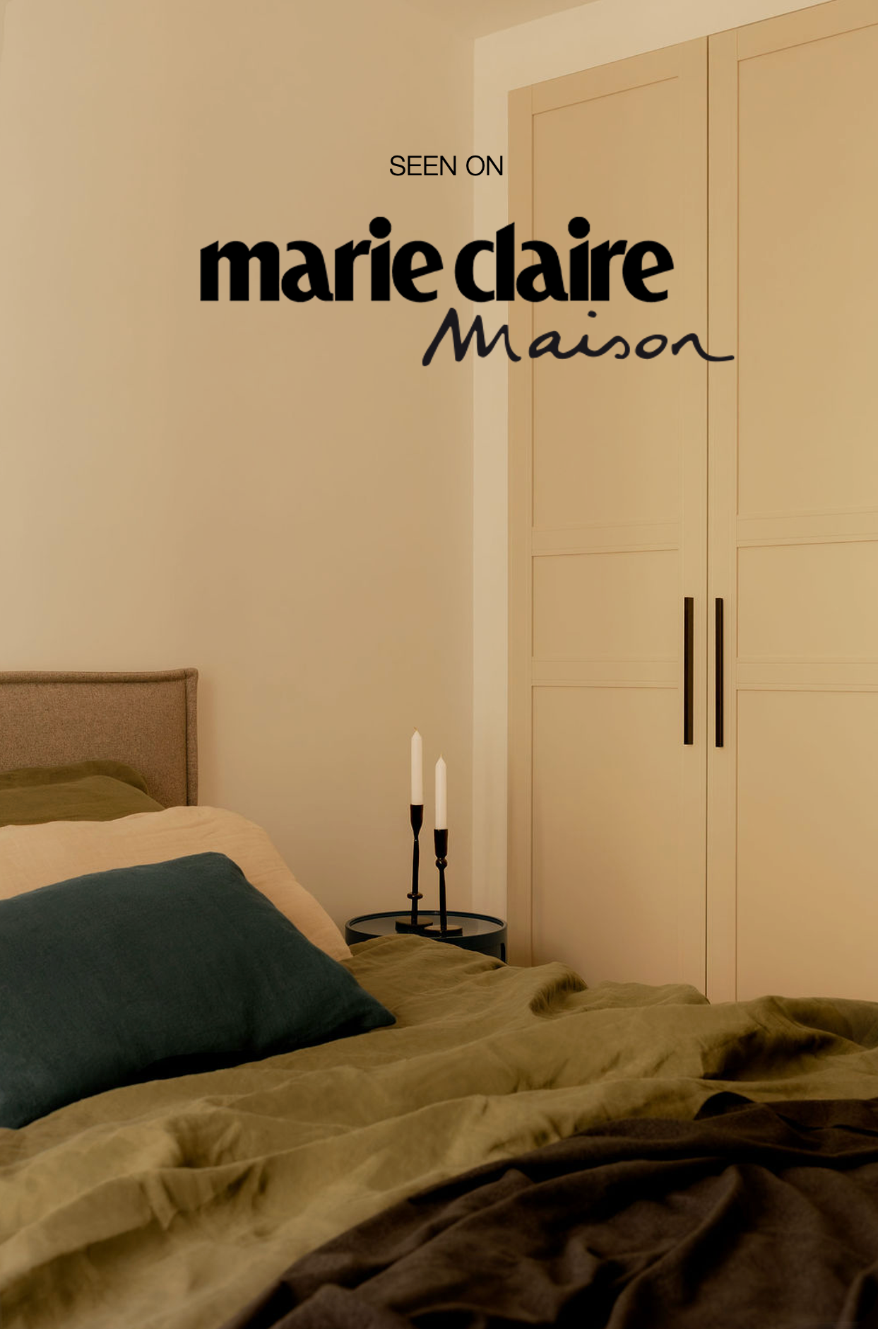 Chambre avec lit fait, coussins, une armoire blanche avec deux poignées noires, une petite table ronde noire avec deux bougies blanches, et un mur beige clair.