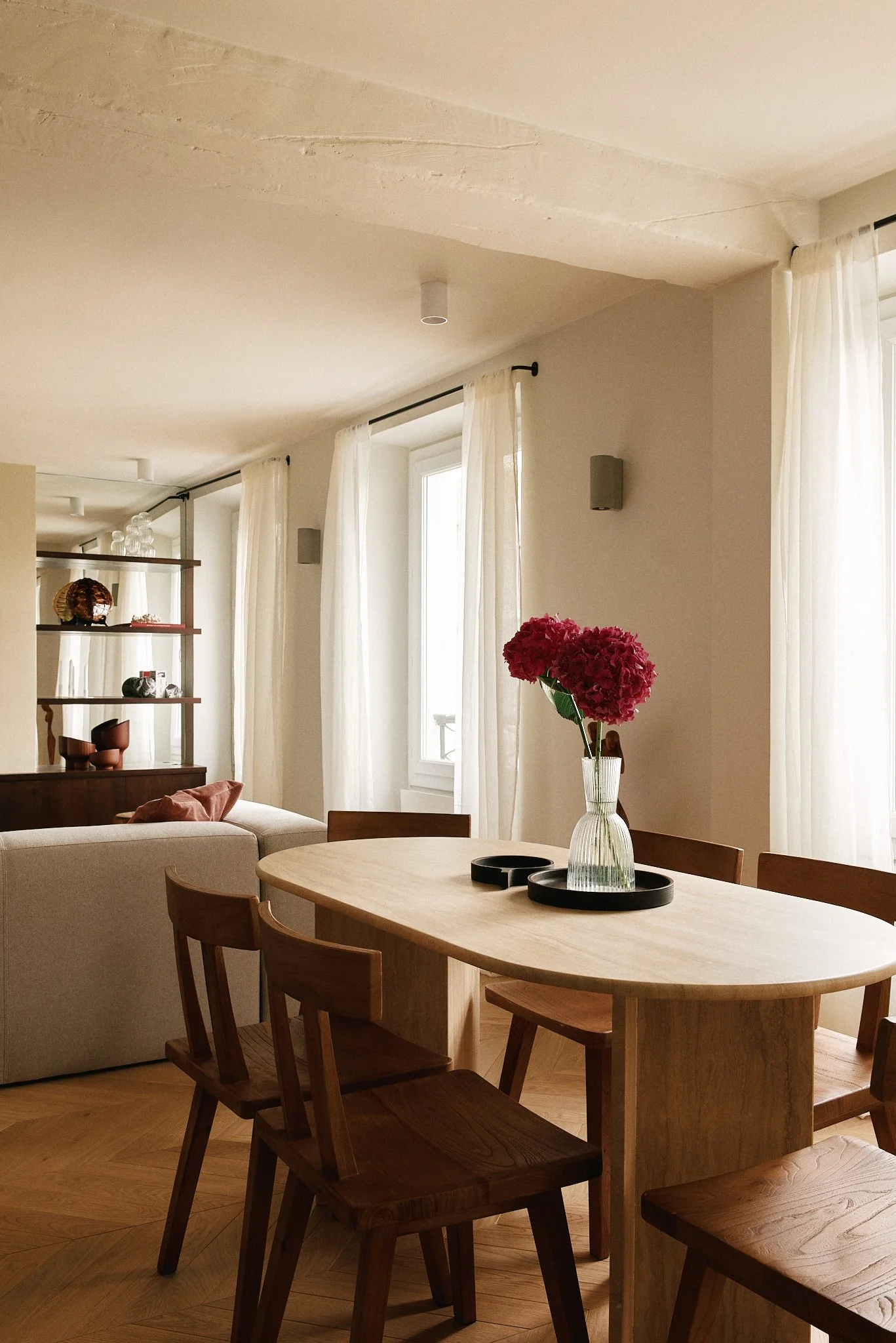 Salon et salle à manger avec table en bois, vase avec des hortensias roses, chaises en bois, fenêtres avec rideaux blancs, étagère avec objets décoratifs.