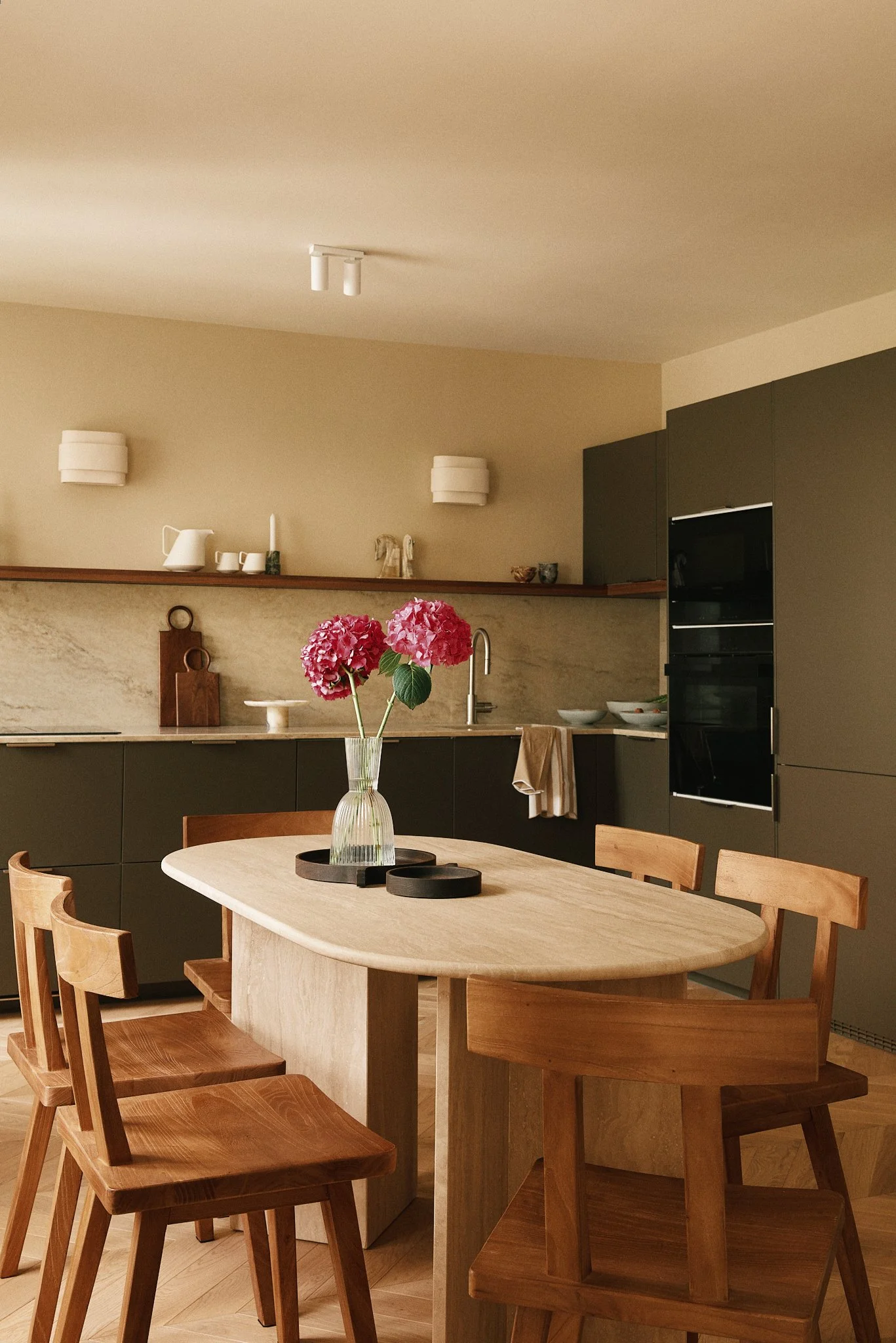 Une cuisine moderne avec un plan de travail en marbre, des armoires noires, un évier en acier inoxydable, et une table en bois avec des chaises en bois clair. Au centre, une vase avec des hortensias roses.