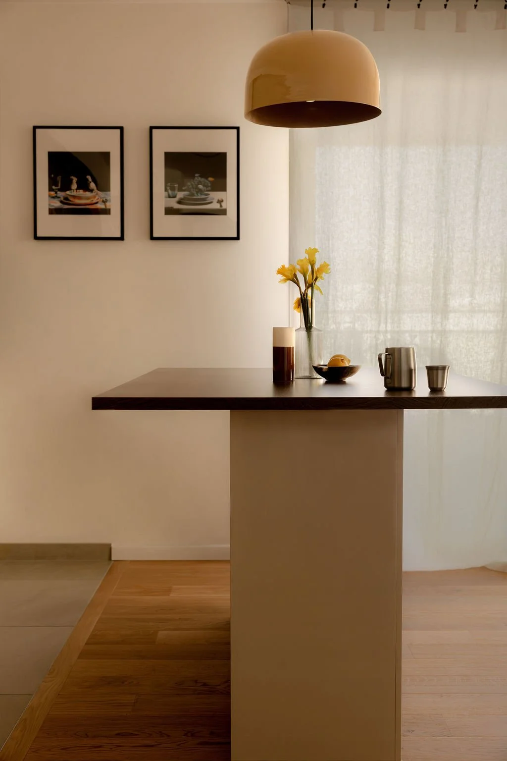 Intérieur d'une salle à manger ou cuisine avec un plan de travail en bois clair, décorée avec un vase de fleurs jaunes, des tasses, une cruche, une coupe avec des fruits, et un abajur suspendu beige.