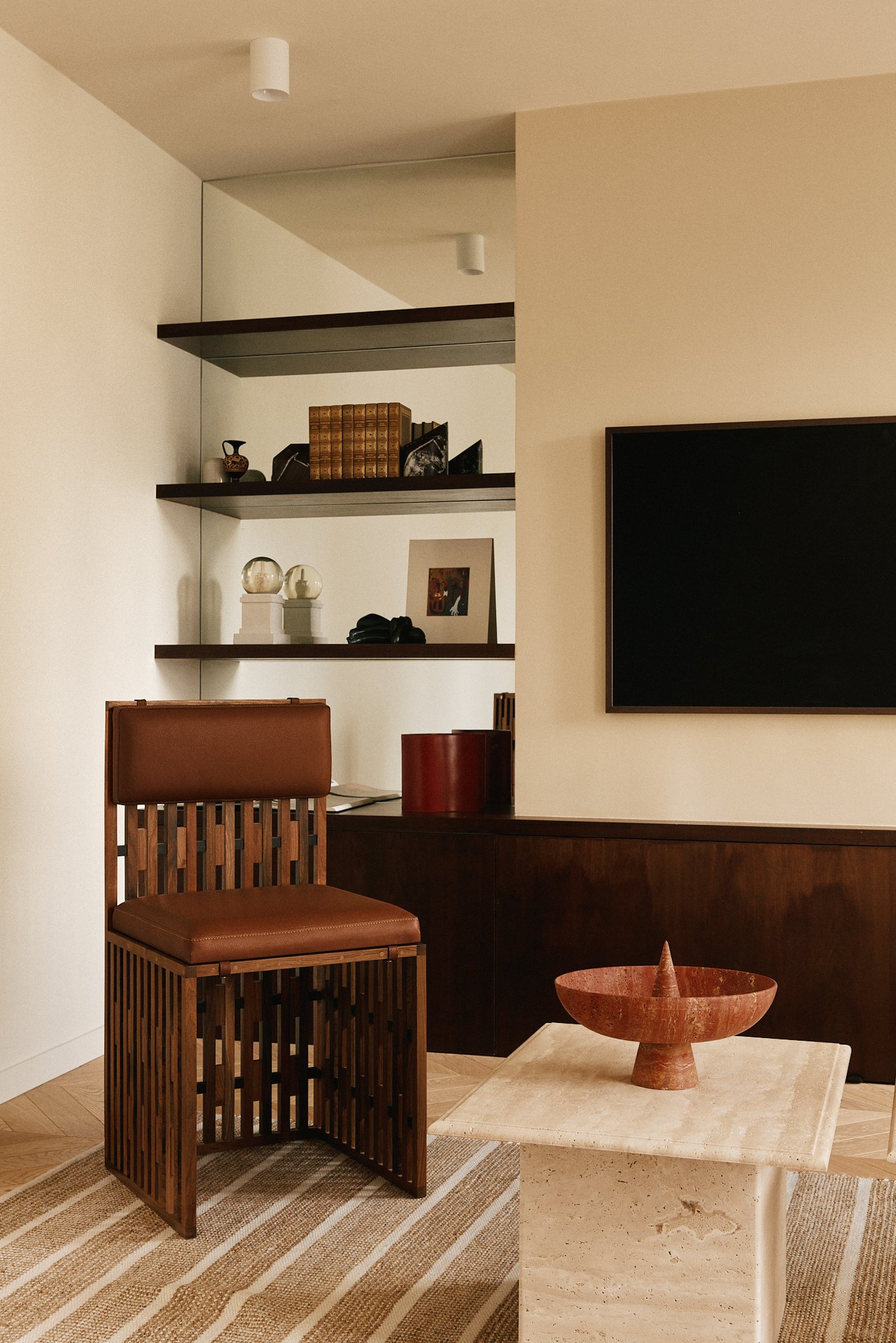 Intérieur d’un salon avec une chaise en bois, une table en pierre avec un bol en terre cuite, et des étagères noires avec des objets décoratifs, des livres, et une télévision à écran noir.
