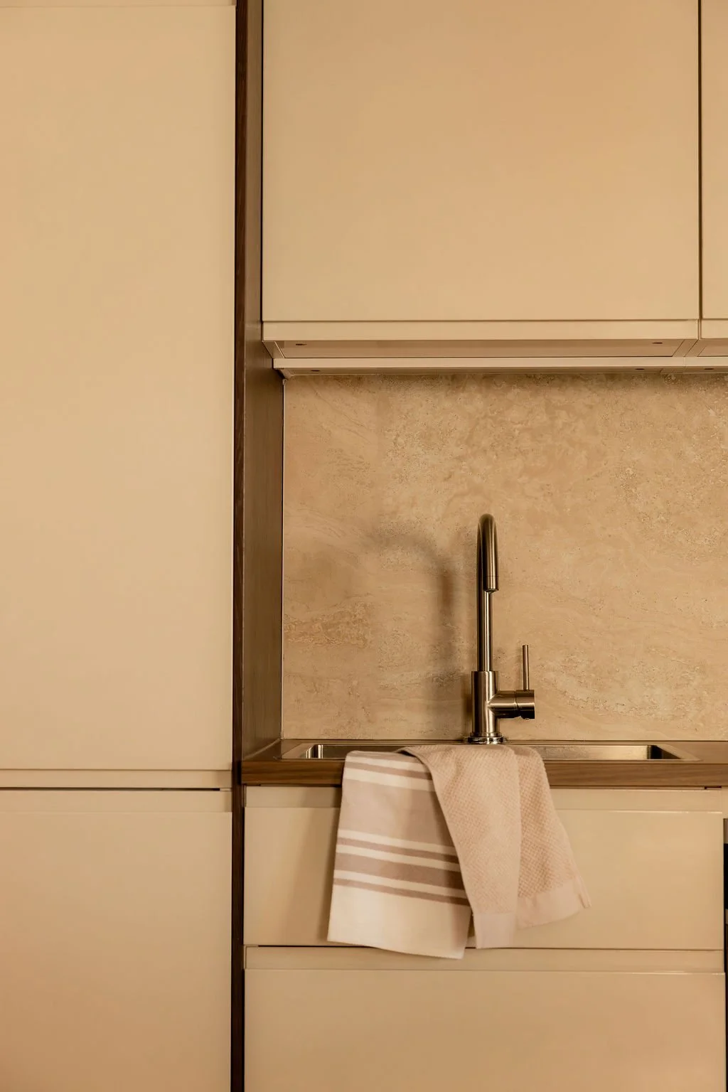 Évier de cuisine avec un robinet en métal, un linge beige suspendu au bord de l'évier, arrière-plan en carrelage beige clair et meubles de cuisine blanc.