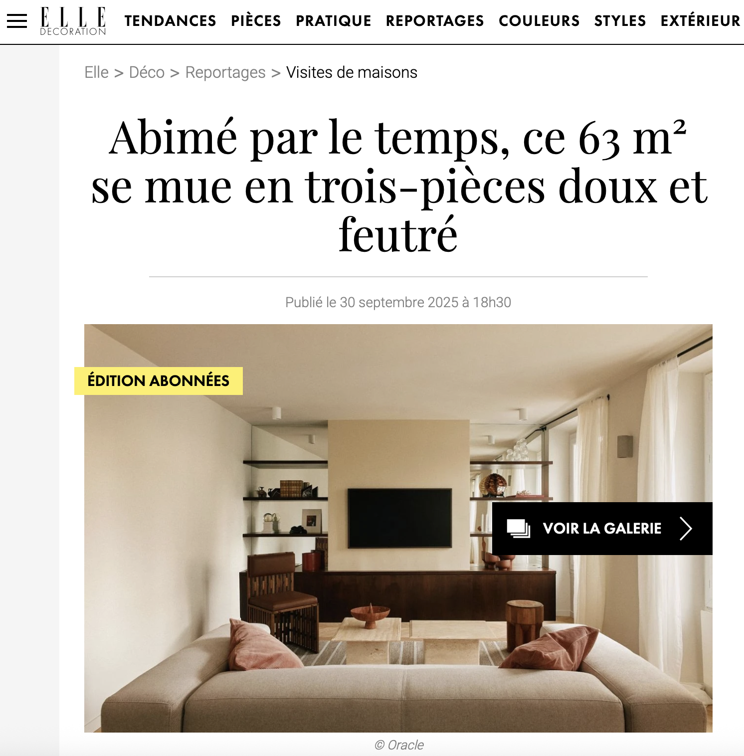 Photo d'un salon moderne avec un canapé beige, des coussins roses, une table basse en pierre avec un bol, une télévision à écran plat sur un mur beige, et des étagères en bois avec des décorations. Fenêtres avec rideaux blancs.