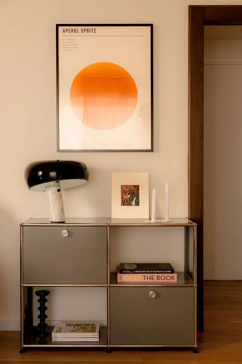 Une étagère en métal gris avec trois compartiments, décorée avec une lampe noire et blanche en forme de champignon, un cadre avec une œuvre d'art, deux bougies blanches, un livre intitulé "IDEAT THE BOOK" et une pile de magazines, contre un mur avec 