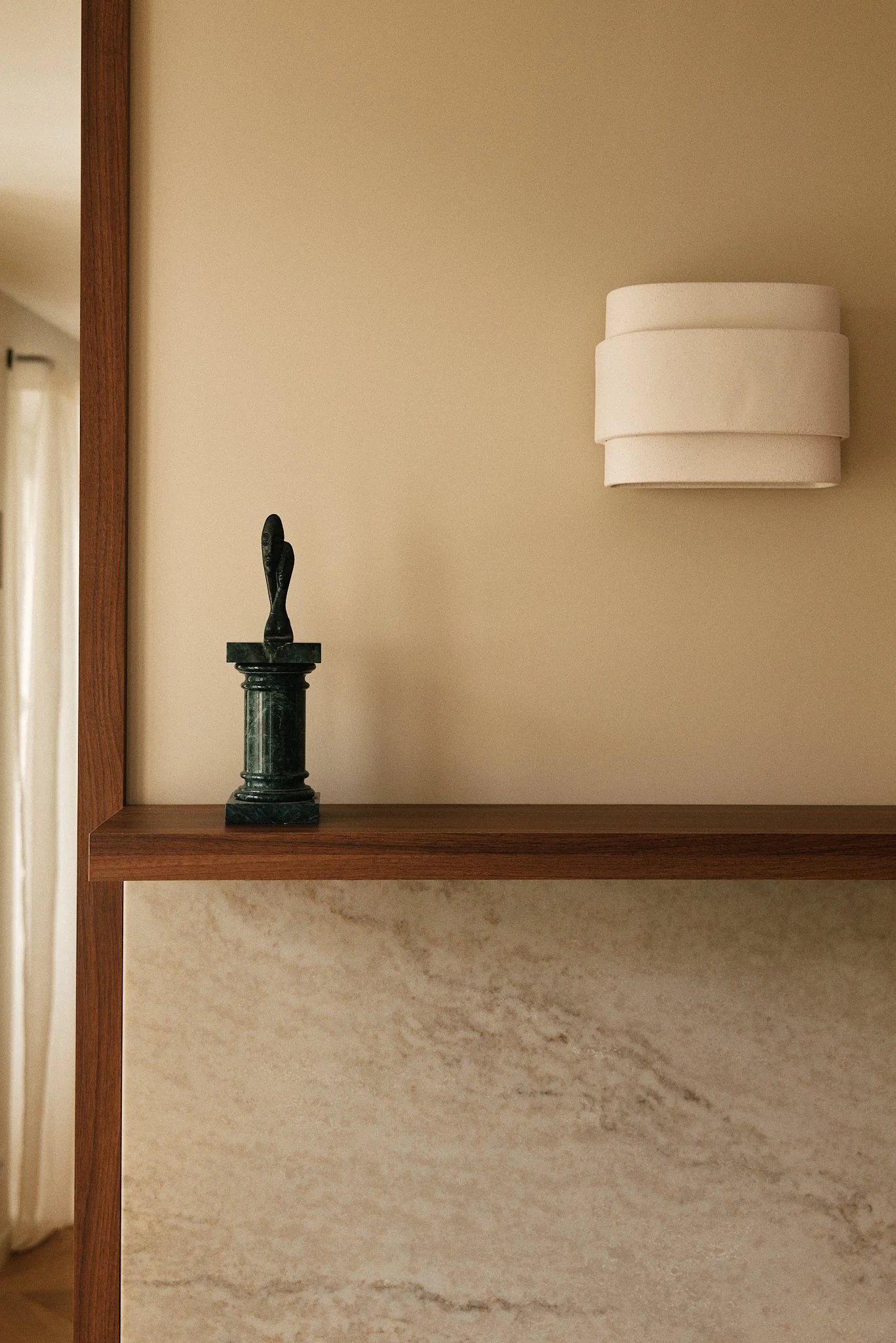 Une étagère en bois avec une petite sculpture en pierre noire en forme de figurine humaine et une lampe en forme de cylindre blanc à plusieurs couches.