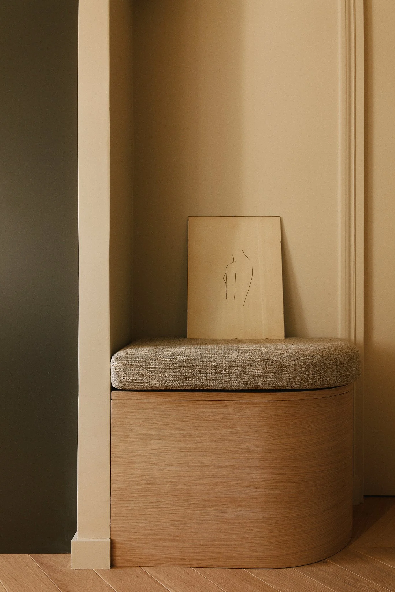 Un coin salon avec un banc en bois courbé recouvert d’un coussin gris, une toile avec une dessin minimaliste d’un corps humain en guise de décoration, dans une pièce avec des murs beige et un sol en bois.