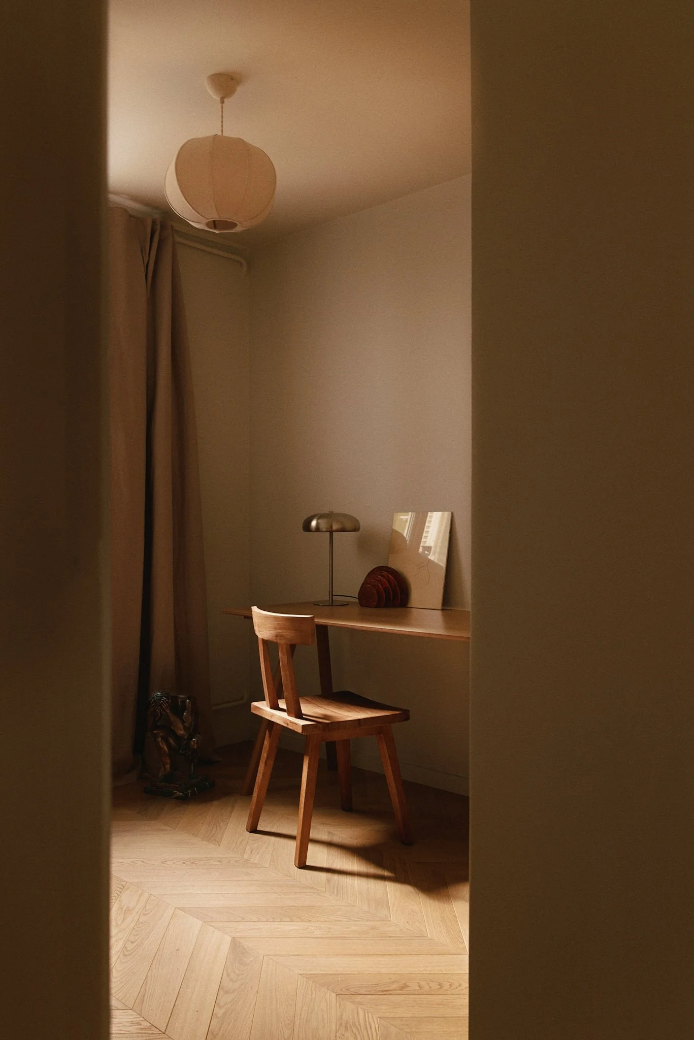 Une pièce avec un bureau en bois, une chaise, une lampe de bureau, un miroir et une sculpture abstraite, avec un lit ou un canapé en arrière-plan et des rideaux beige.