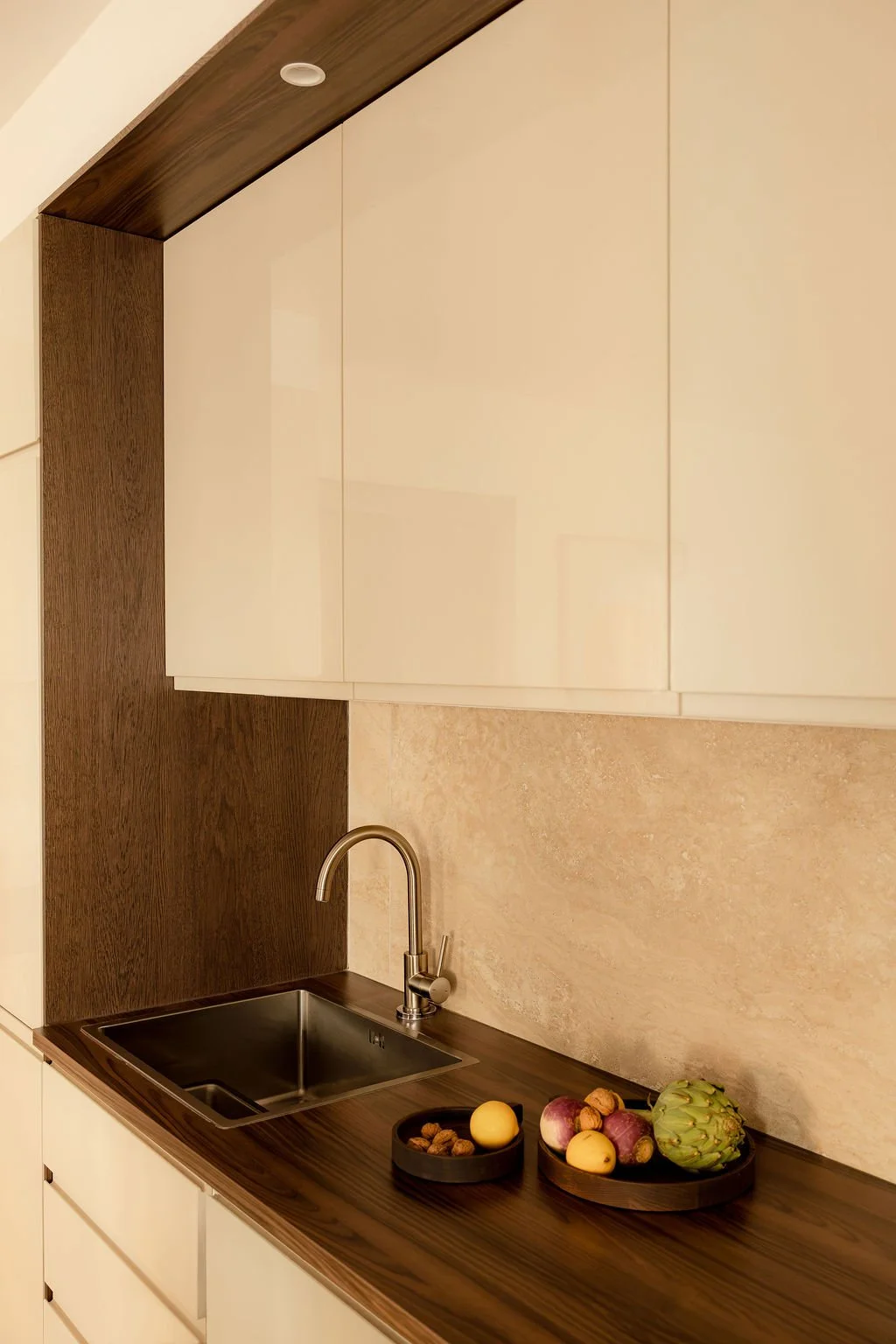 Une cuisine moderne avec un évier en acier inoxydable, un robinet en métal, des plans de travail en bois foncé, et des fruits disposés dans des paniers.