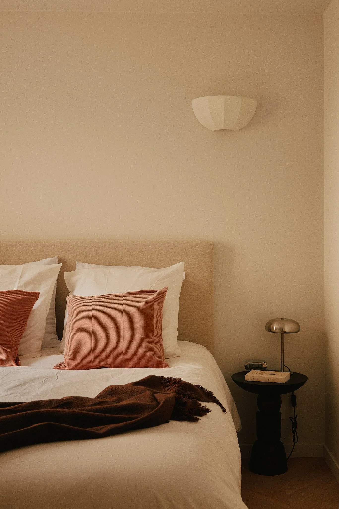 Chambre avec lit à la tête en tissu beige, oreillers blancs et coussins roses. Lampe de chevet sur une table noire, avec livre et radio.