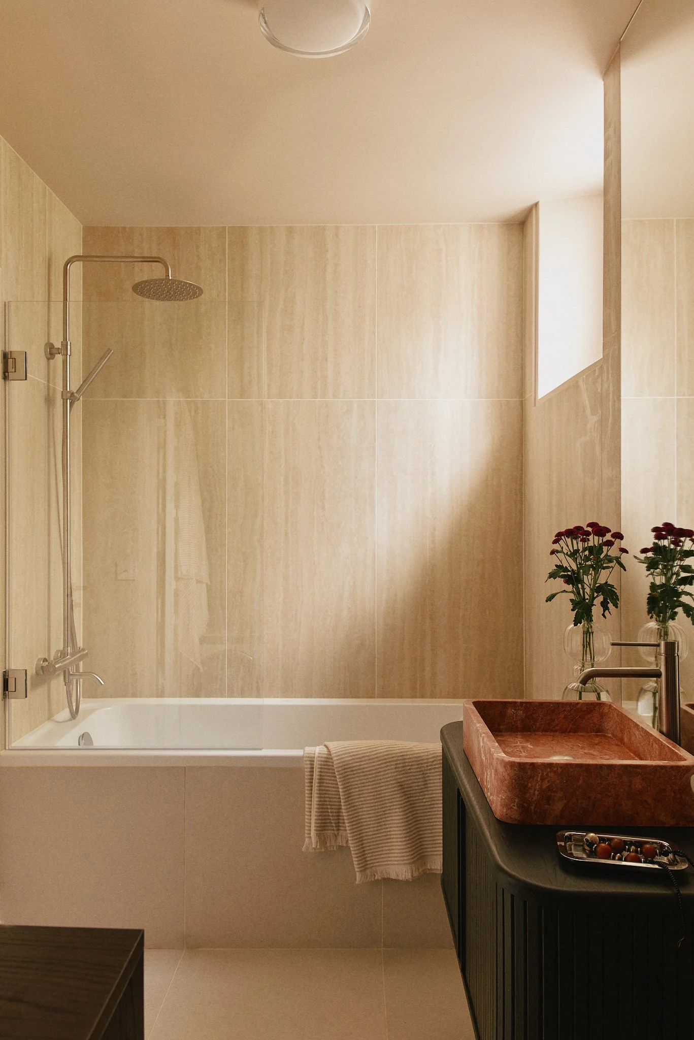 Salle de bain avec baignoire,paroi en verre, douche à main, mur en carrelage beige, lavabo en pierre rouge, meuble noir, deux vases avec des fleurs rouges, et un miroir.