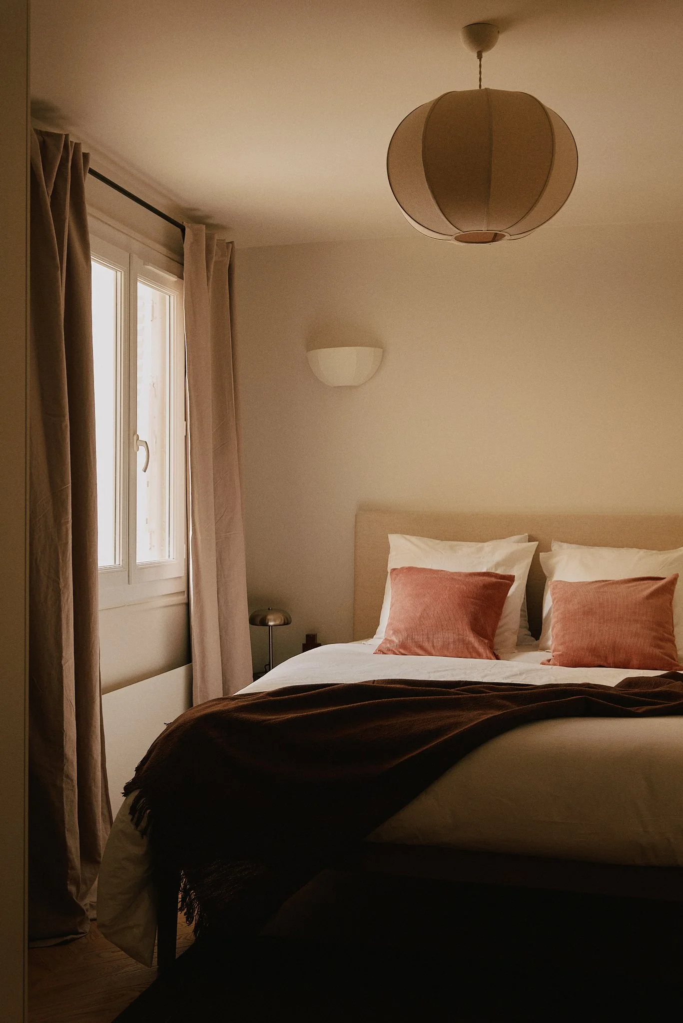 Chambre à coucher avec un lit fait, draps blancs, coussins rose sur le lit, fenêtre avec rideaux beige, lampe de chevet à gauche, plafonnier beige, mur blanc.