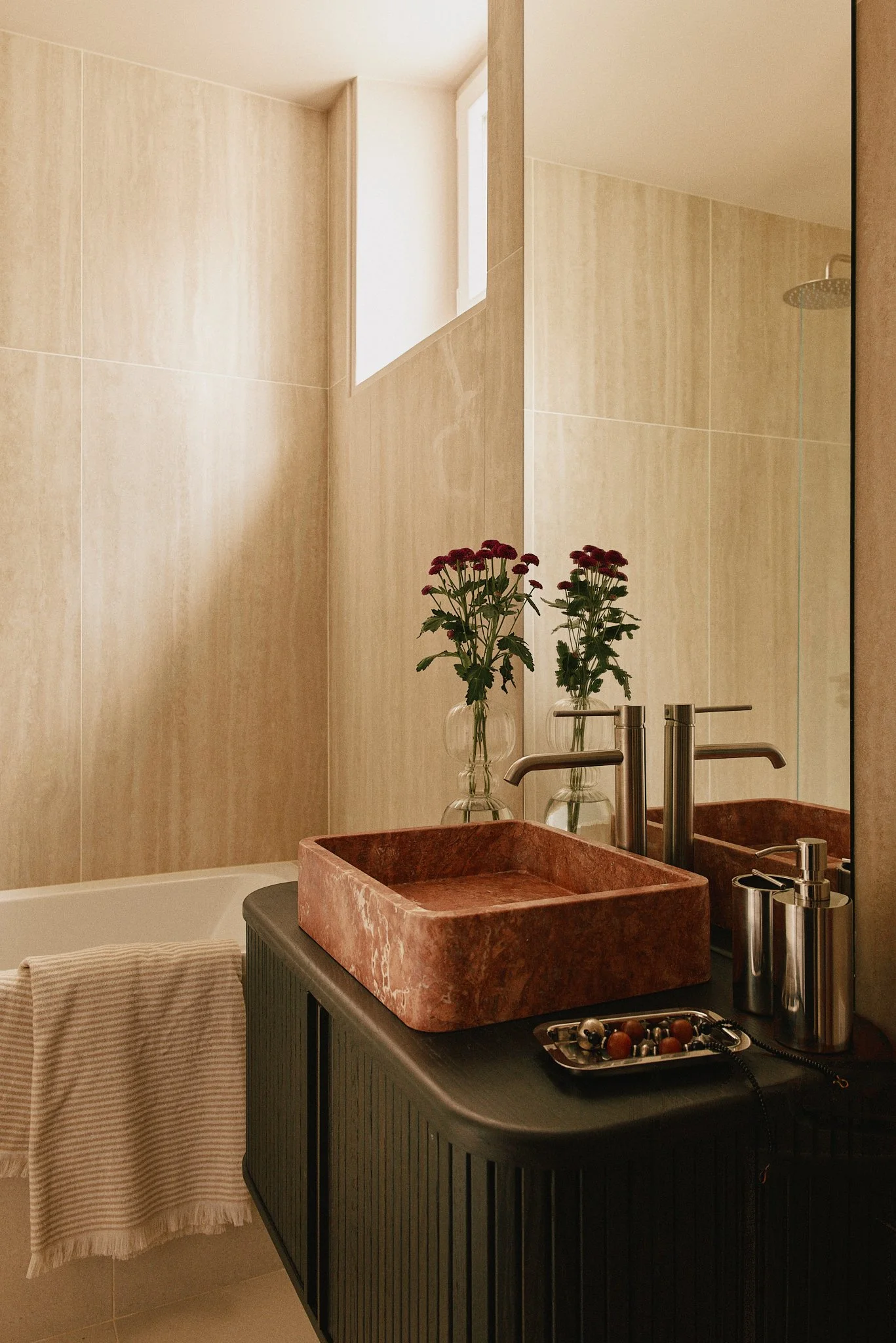 Salle de bain avec un lavabo en pierre de couleur rougeâtre, miroir, fleurs dans des vases en verre, serviette blanche, et une douche à l'italienne, murs en carrelage beige, au plafond une fenêtre.