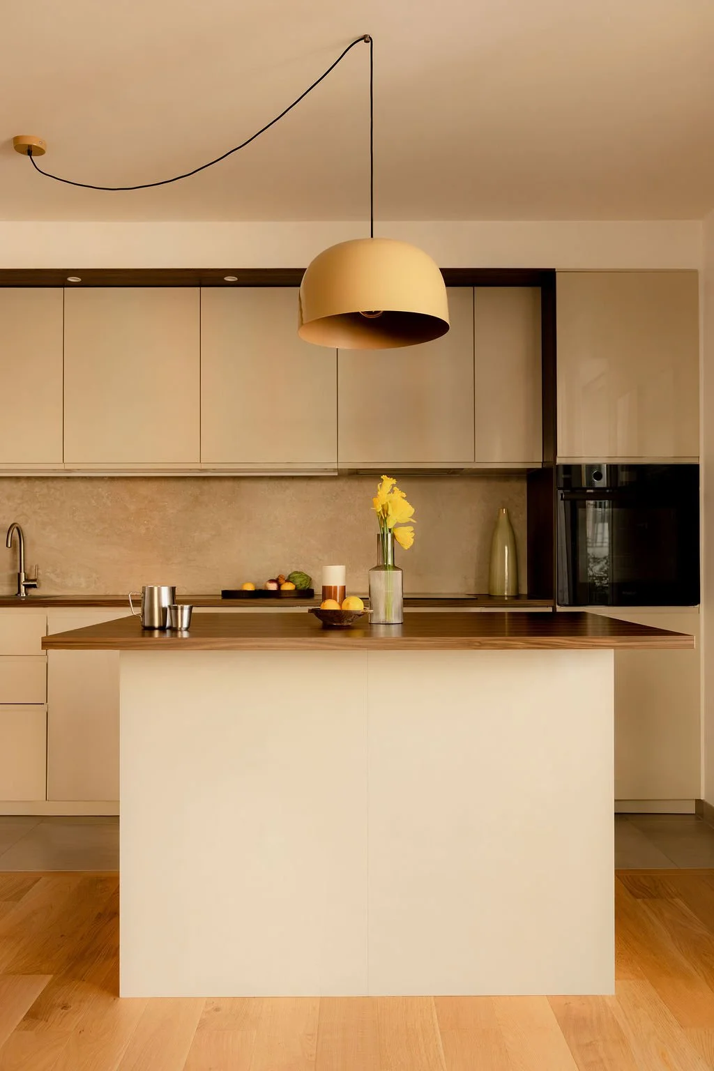 Cuisine moderne avec un plan de travail en bois, un mur beige, des armoires beige, une étagère en bois dans le mur, un évier, une lampe suspendue beige, des fleurs jaunes dans un vase, des fruits et une bouteille avec un verre.