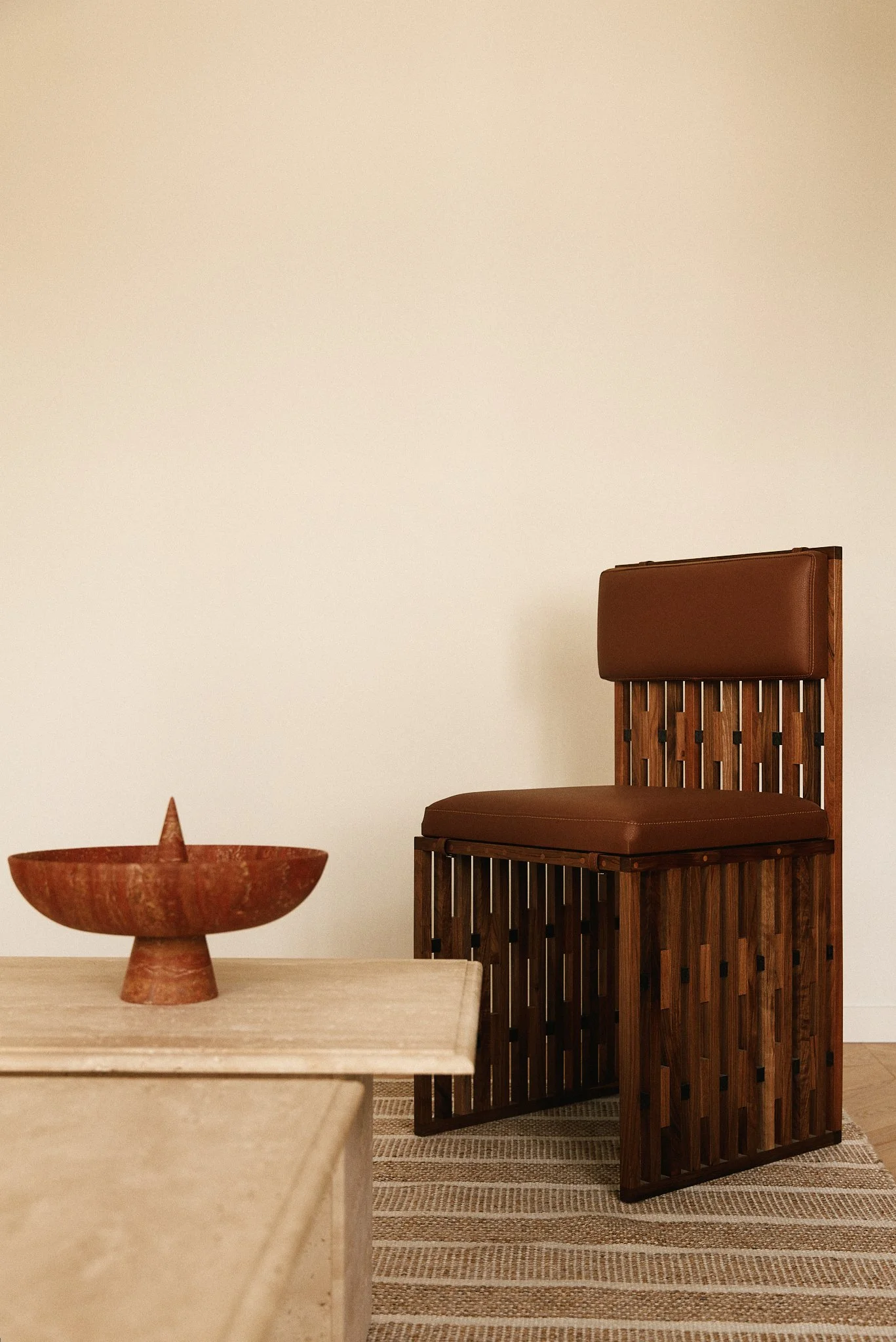 Une chaise en bois avec un coussin en cuir marron et un repose-tête, située à côté d'une table en bois clair avec un bol en pierre rouge dessus. La scène est sur un tapis rayé beige et marron, contre un mur blanc.