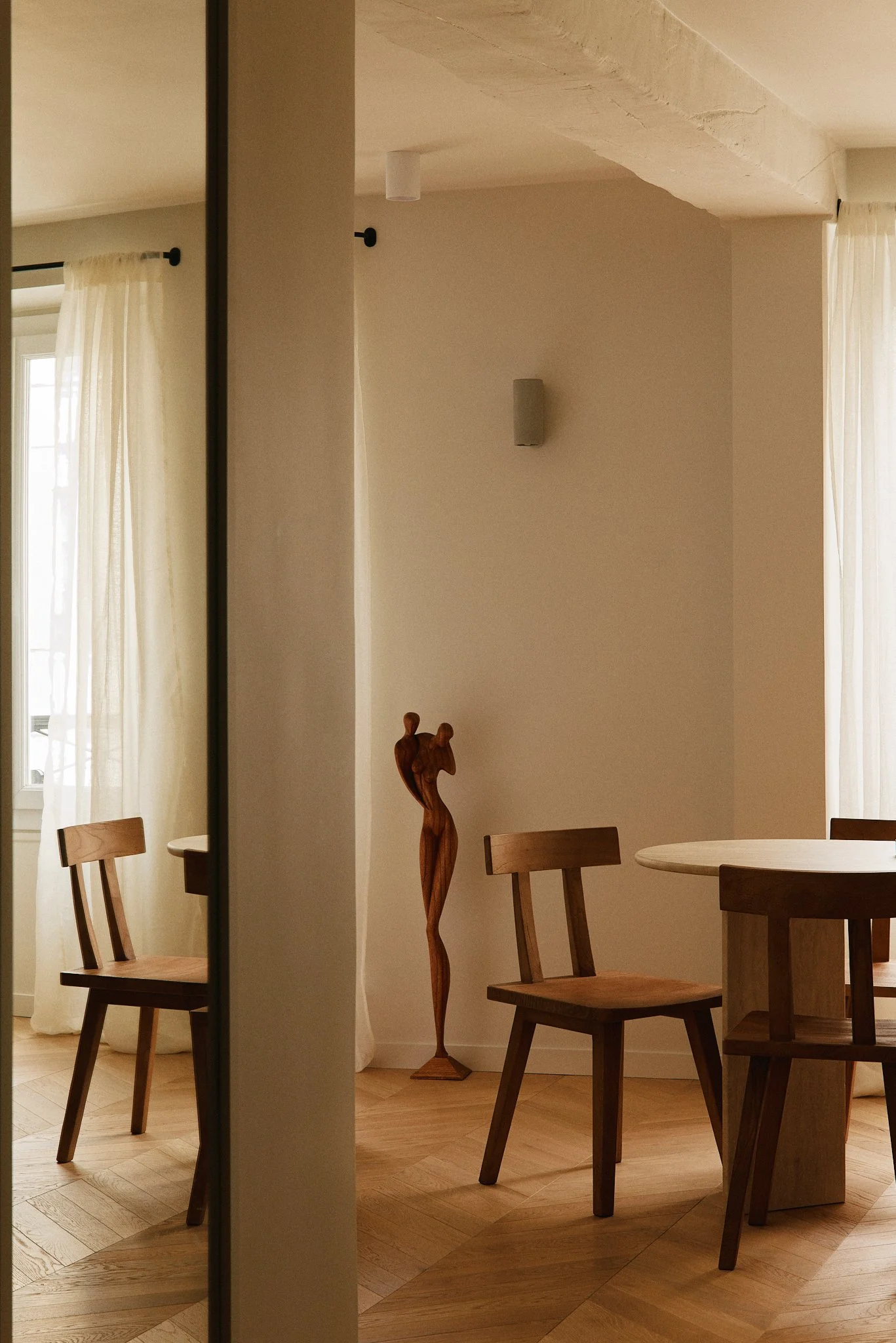 Intérieur d'une pièce avec meubles en bois, rideaux blancs, sculpture en bois, et murs clairs.
