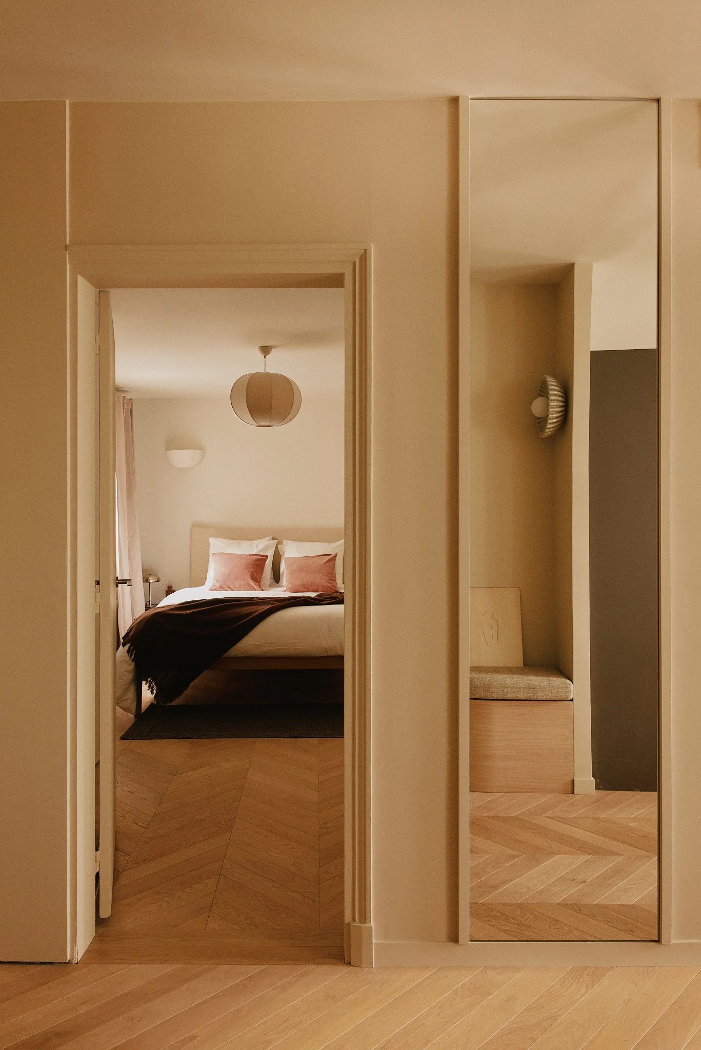 Vue d'une chambre d'hôtel, vue à travers une porte ouverte. La chambre comporte un lit avec des oreillers roses, une suspension en papier, une table de nuit avec une lampe, et des rideaux roses. À droite, un mur avec un miroir et une petite banquette