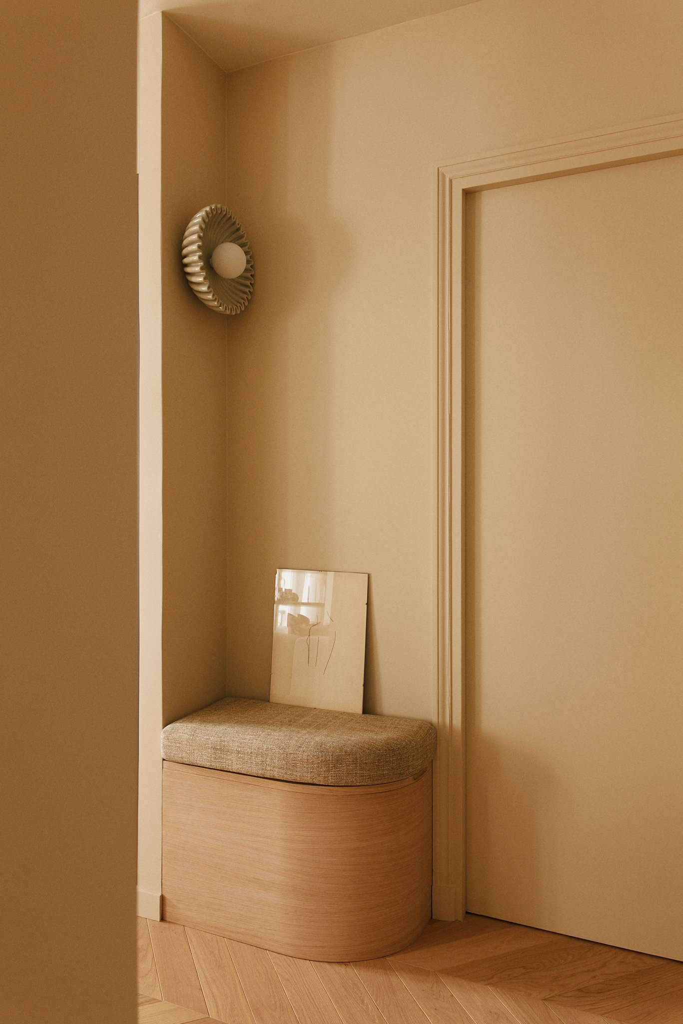 Coin d'entrée avec un banc rembourré en bois clair, un miroir appuyé contre le mur et une lampe murale décorative en forme de cercle avec une ampoule blanche, dans une pièce à murs beige clair.